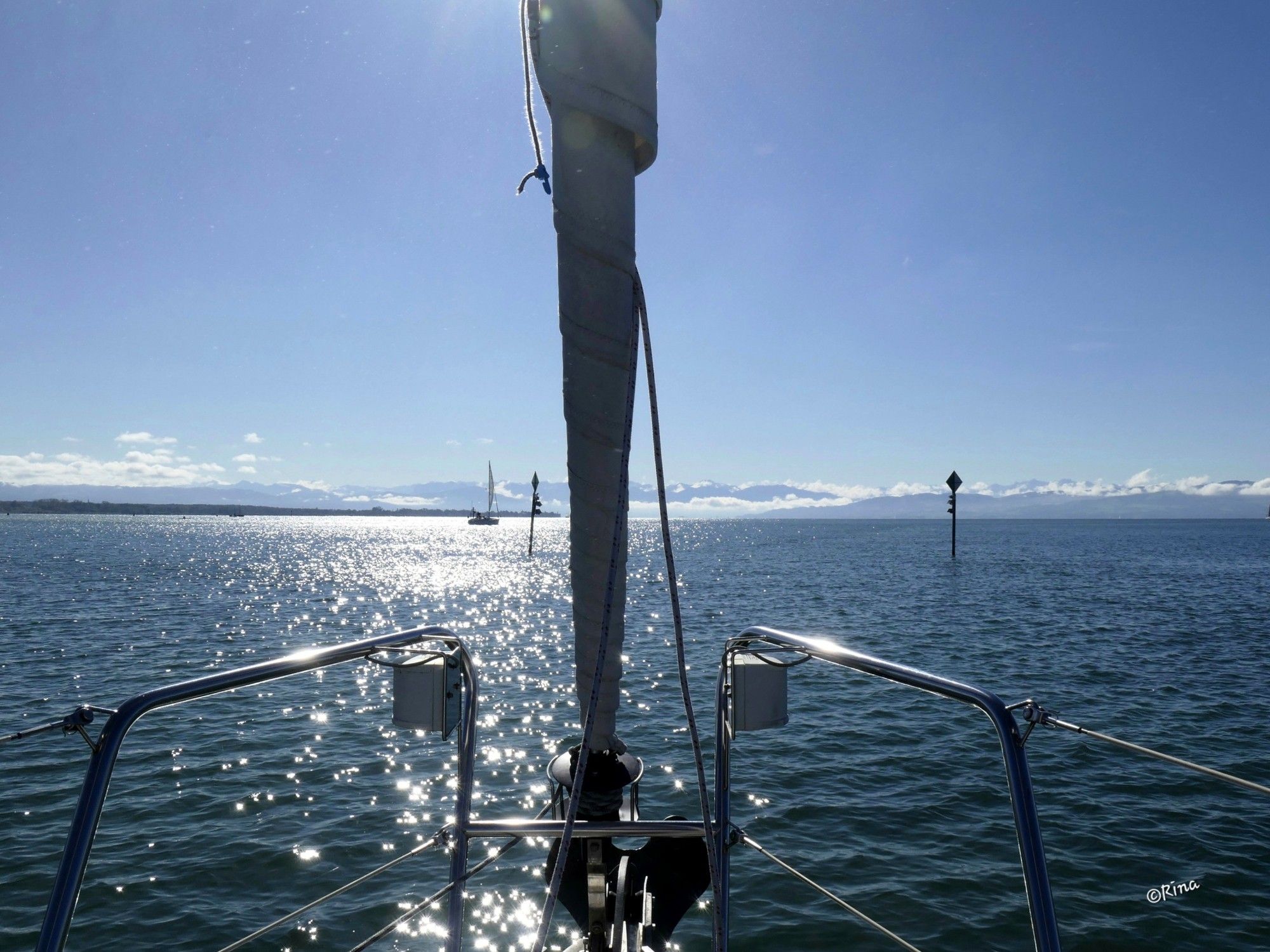 Man sieht den Bug eines Schiffes. Die Sonne spiegelt sich so auf dem Wasser, so das es wie Sterne glitzert.  Im Hintergrund sieht man Wolken, die tief in den Alpen hängen, die Gipfel sind aber frei. Der Himmel ist strahlend blau.