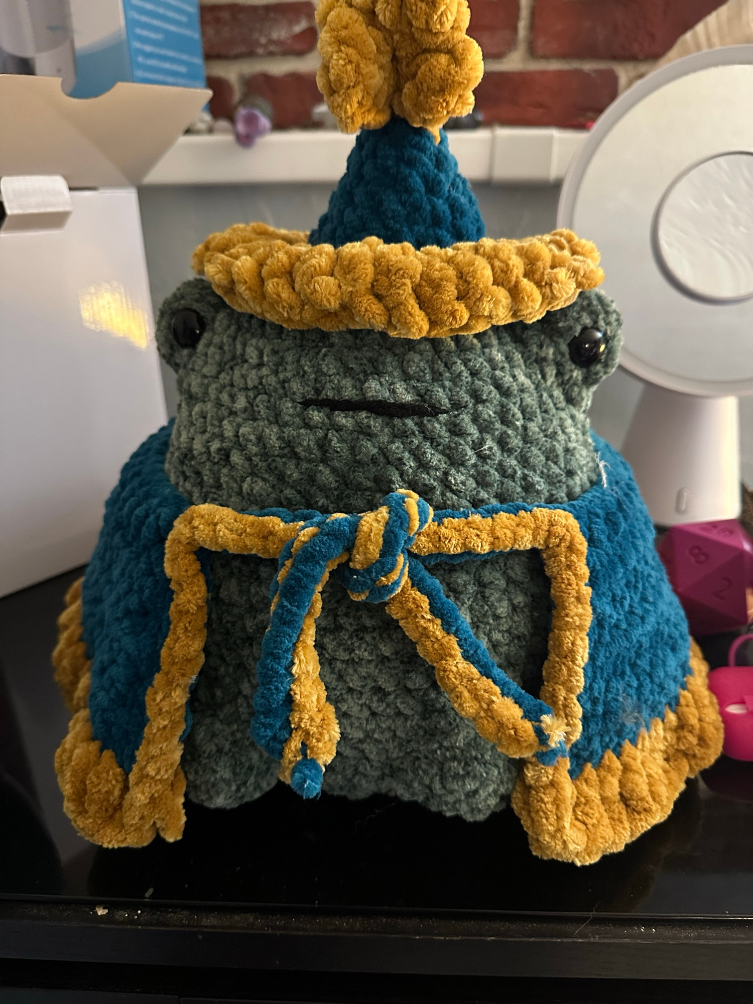 a large crochet frog made with chenille yarn. he has a little blue hat with a gold star on the top and a blue cape with a long gold trim