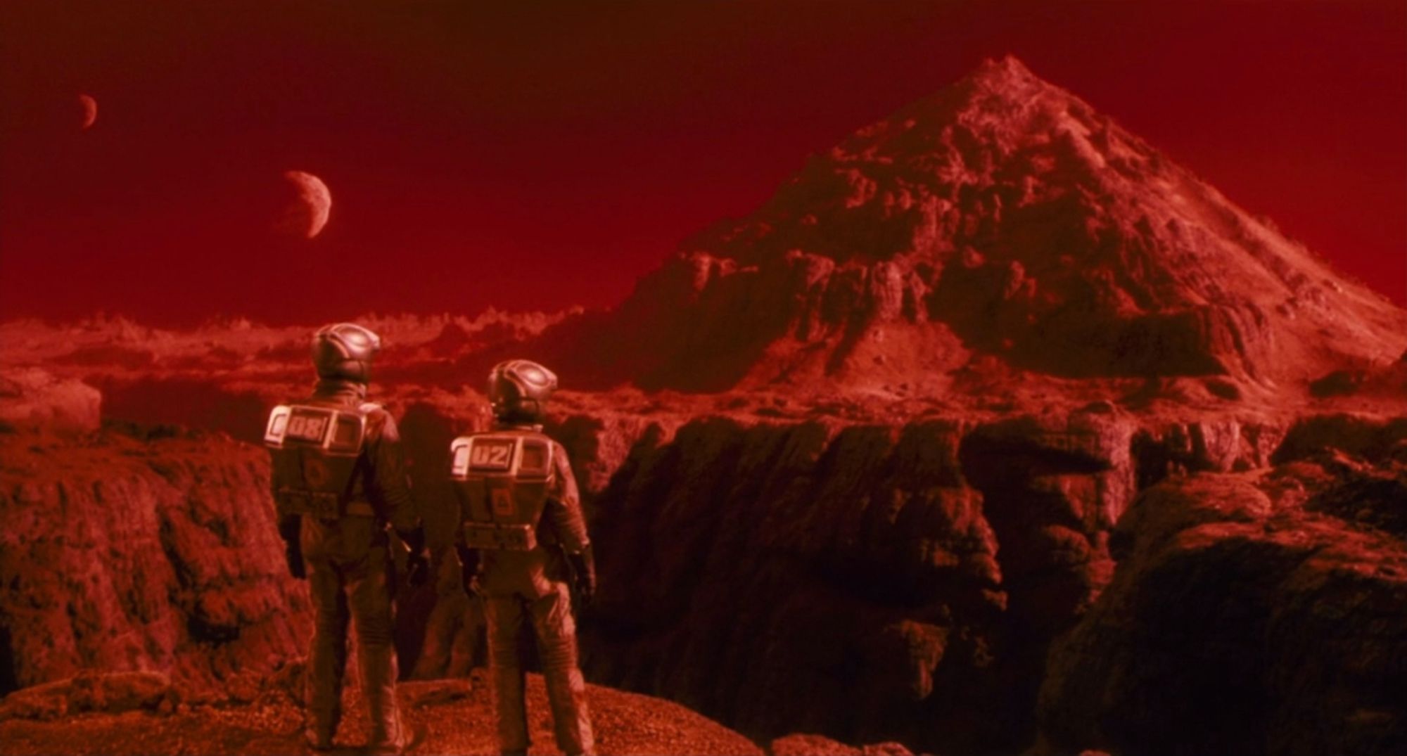 Red rocky Mars canyon landscape with two people in vaccsuits in the foreground, a huge mountain and two moons in the distance

from TOTAL RECALL (1990)