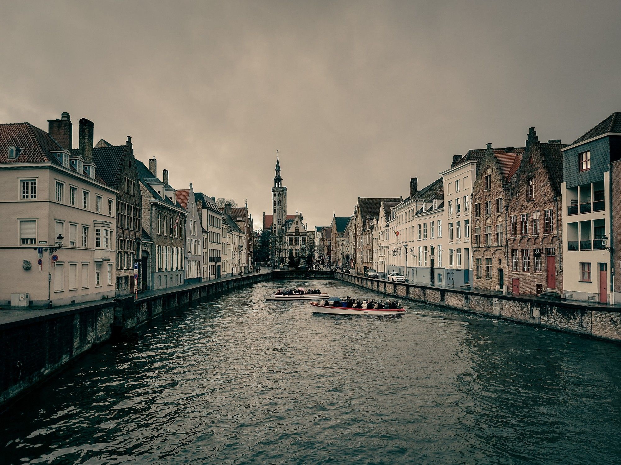 View of a Bruges canal, lined with old houses, showing a spire in the background and two boats.