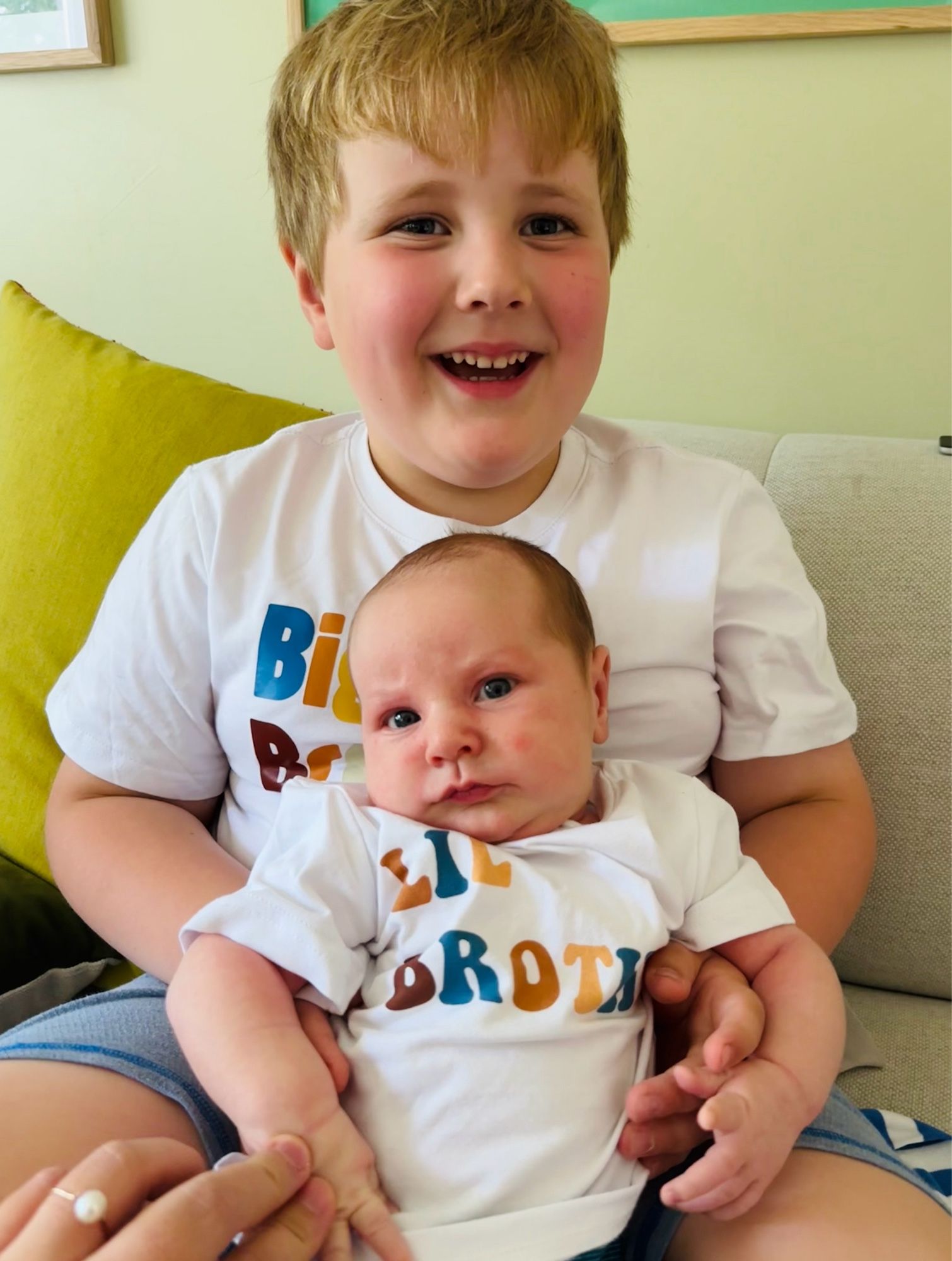 Big brother smiling while holding a non-plussed newborn on his lap. Both wearing matching big bro, lil bro tees.