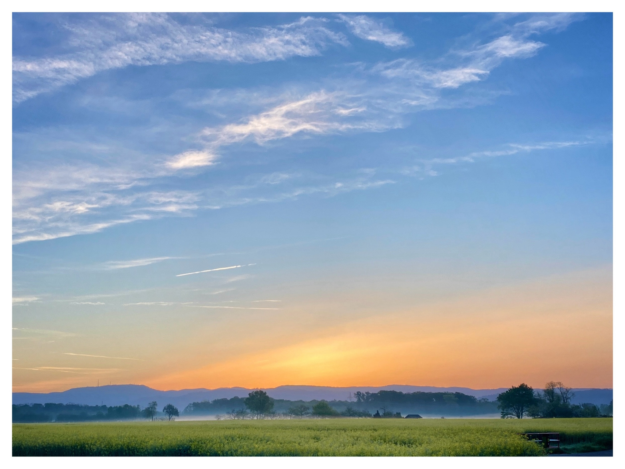 Landschaftsaufnahme, bei der man im Vordergrund ein Stück Wiese sieht. Ein Stück weiter hinten, Nebelschwaden, dann folgen Bäume und dahinter bewaldete Berge. Und hinter dem ganzen Szenario geht die Sonne auf - orange verlaufend in dem blauen Himmel über.