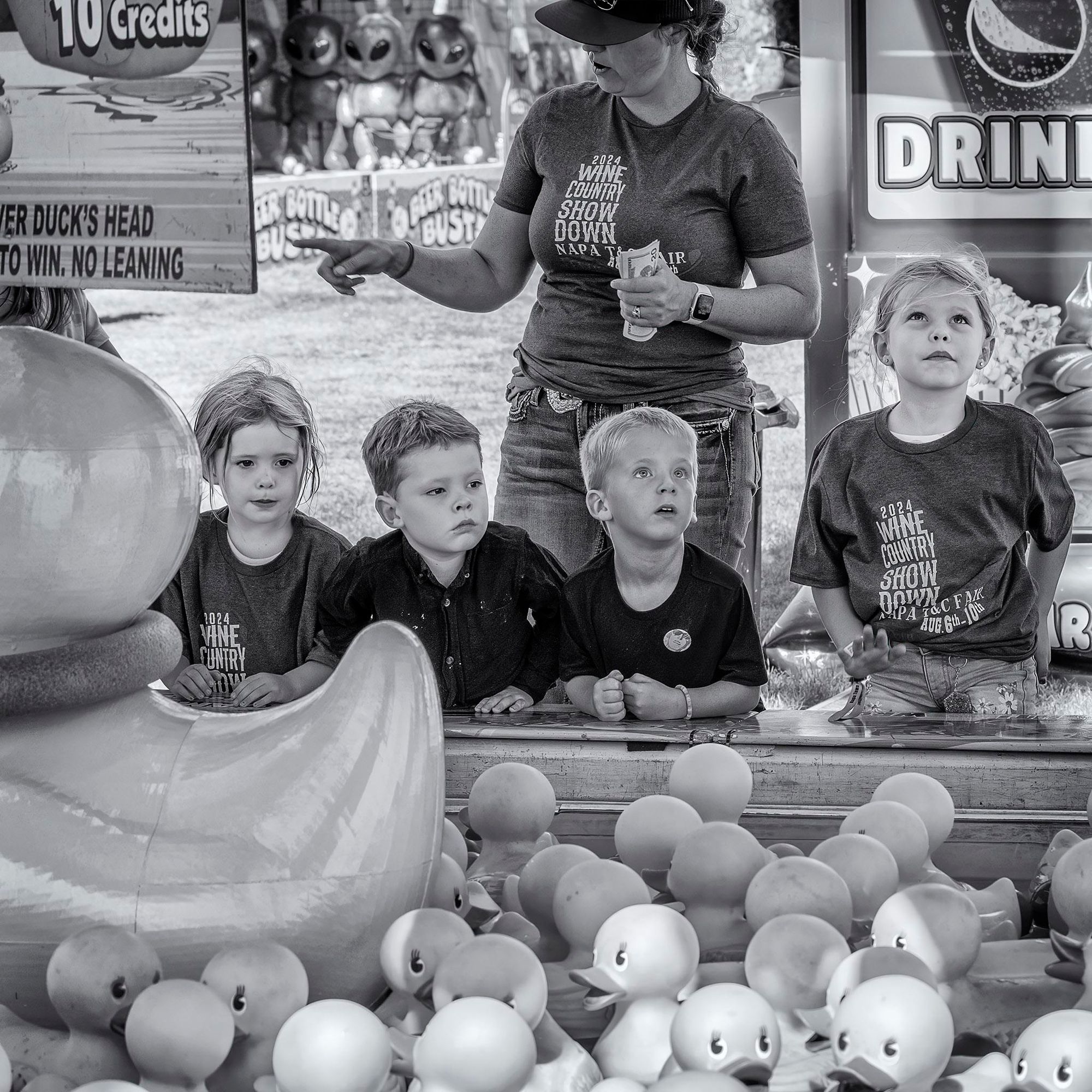 Group of children at the edge of "Ring a Duck" carnival game, which consists of a small pool with rubber ducks floating on it. The goal is to toss a ring over the neck of one of them.