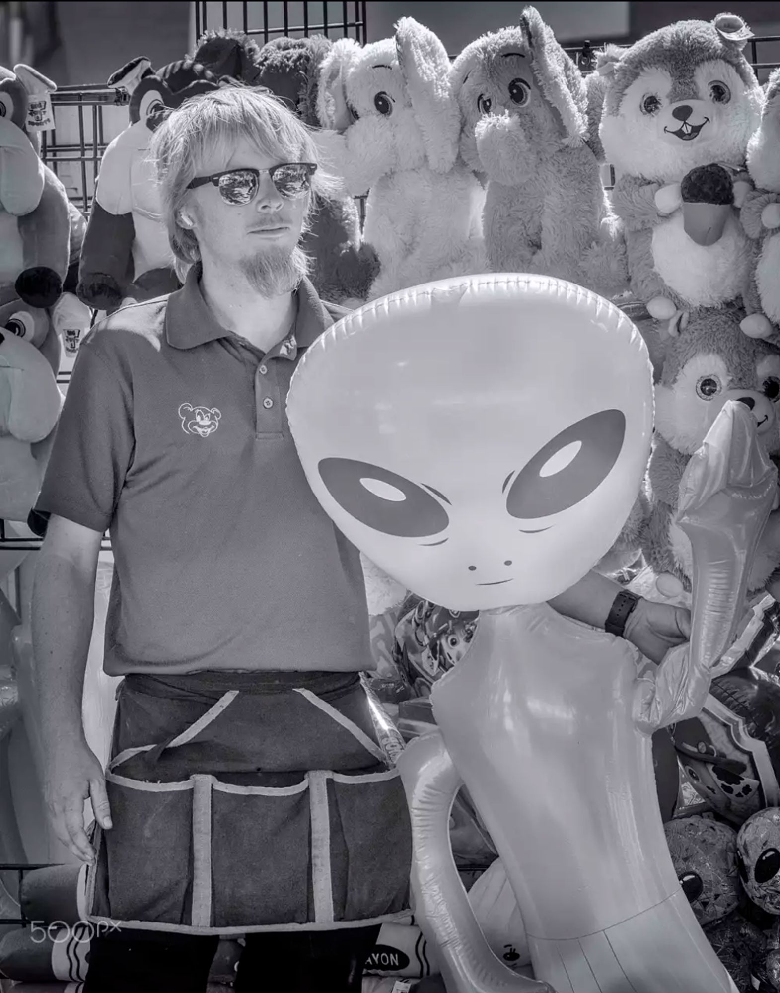 Carnival game operator holding up a prize--a blow up doll made to look like an alien from outer space--in front of a shelf with other prizes including stuffed toys 