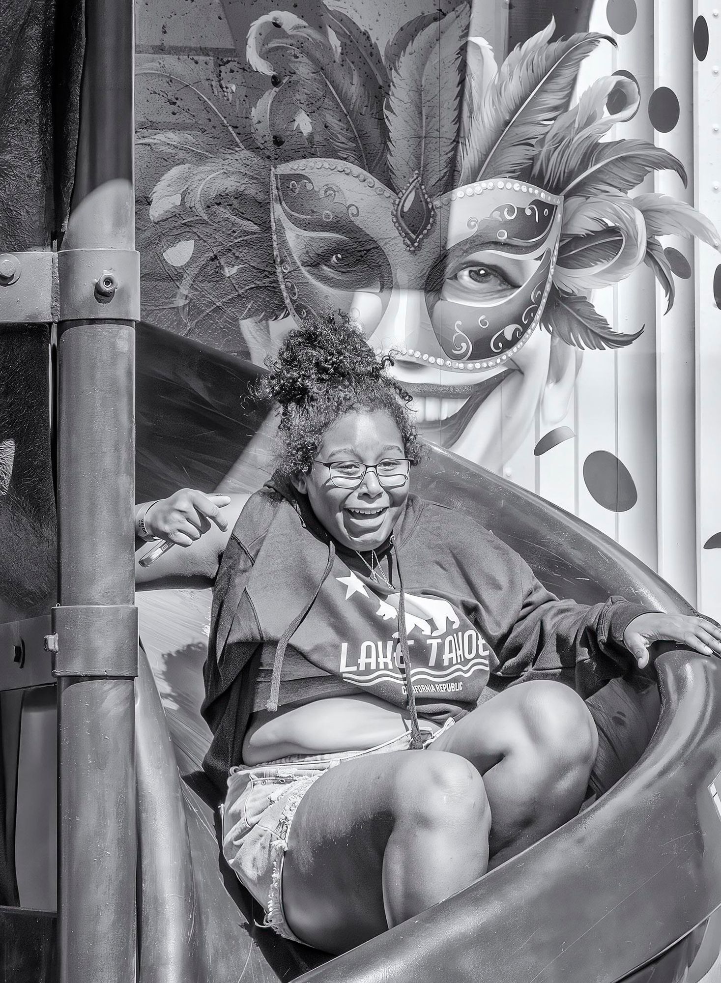 Girl slides down a twisting slide at the end of a "funhouse" ride at the Napa County Fair. A mardi gras character painted on the wall behind her seems to echo her wide grin.