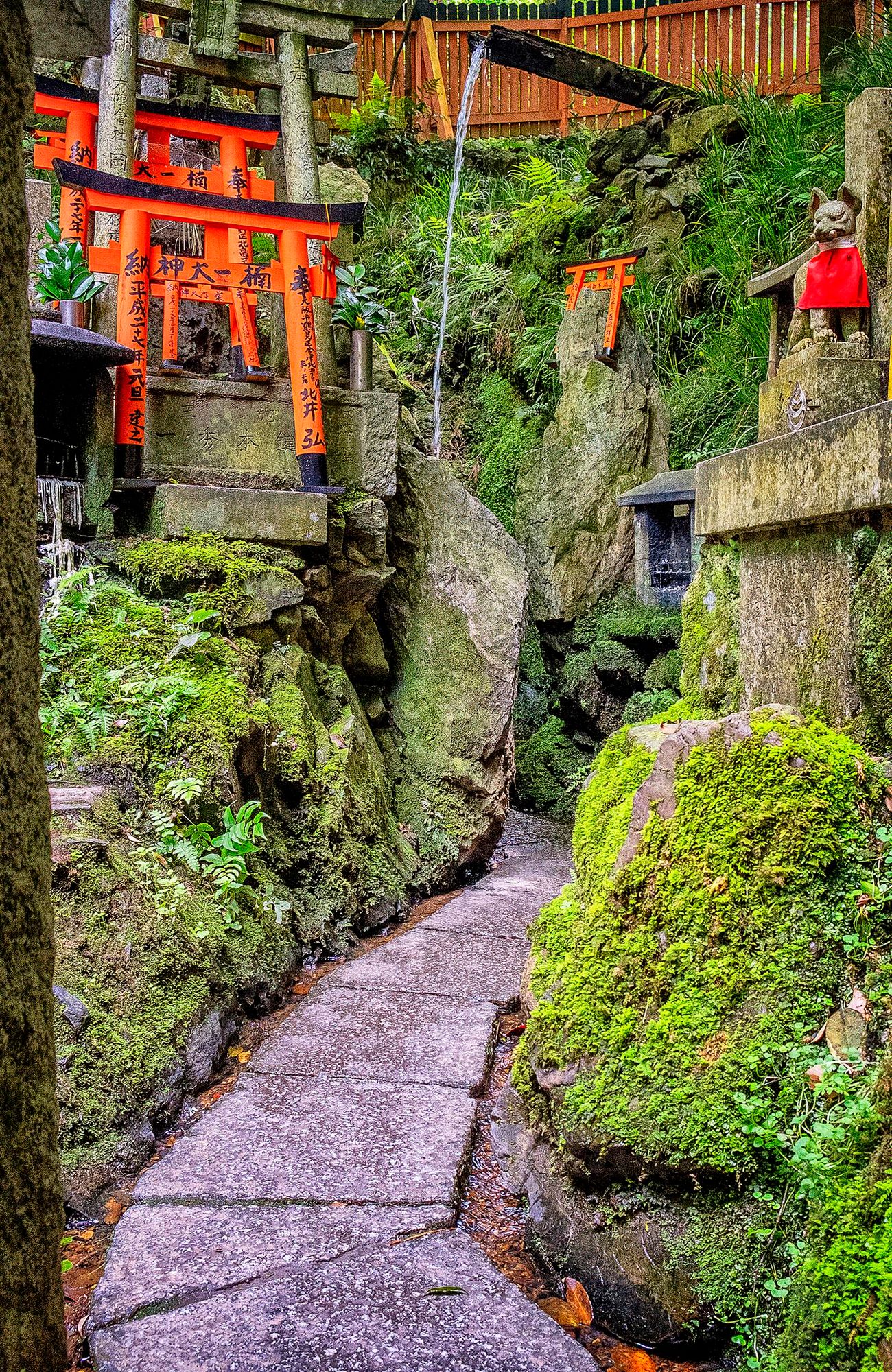 Paved path leading among moss encrusted rocks to a shrine with a fountain and red toriii gates in Kyoto's Fushimi Inari Shrine.