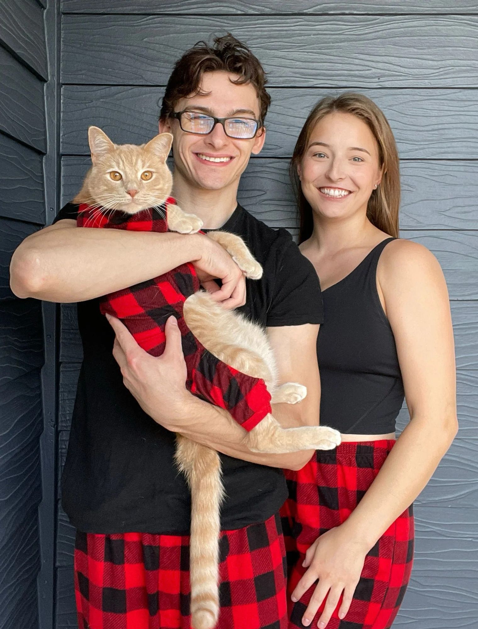 A smiling young couple pose wearing matching black t shirts and red flannel pajama pants, holding an orange tabby cat who is wearing a red flannel cat onesie