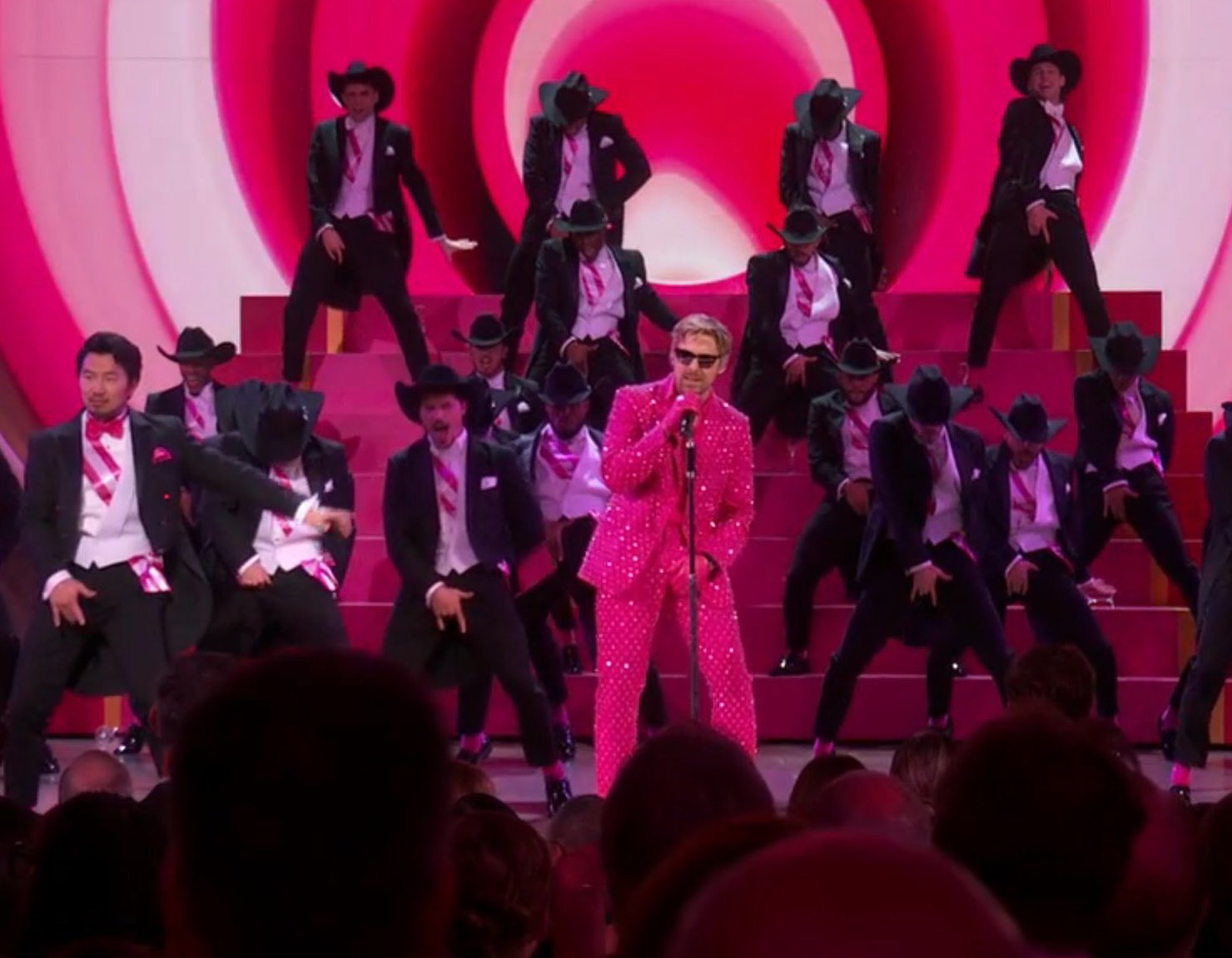 Ryan Gosling and the rest of the Kens from the Barbie movie performing I'm Just Ken at the Oscars. Ryan is dressed in a hot pink rhinestoned suit and the rest of the Kens are dressed in black and white suits with pink ties in reference to the following photo.