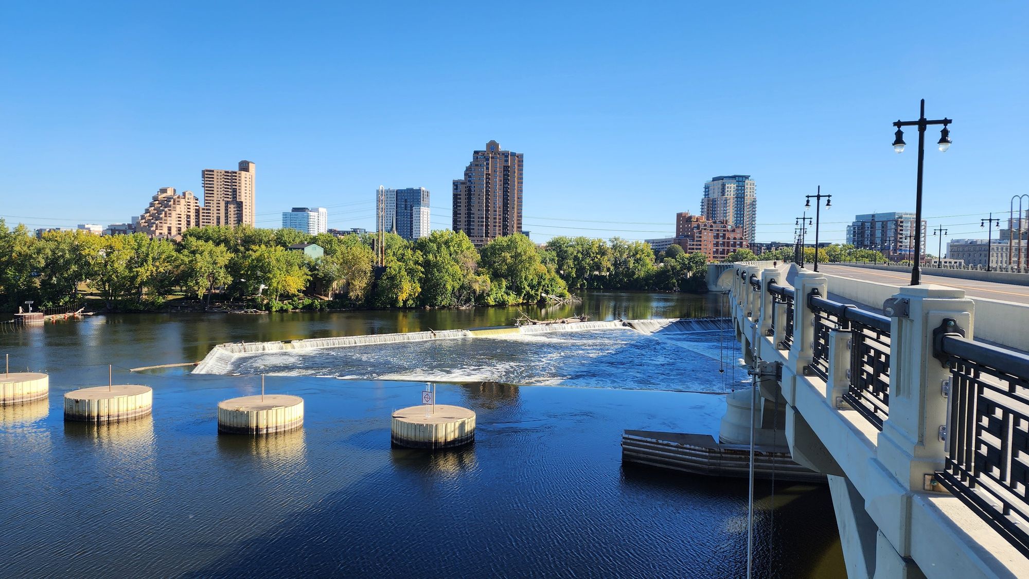 A clear day, blue sky. On the right is a bridge. St Anthony falls creates tubulent water that runs beneath the bridge. Tall residential buildings sprout from the east bank.