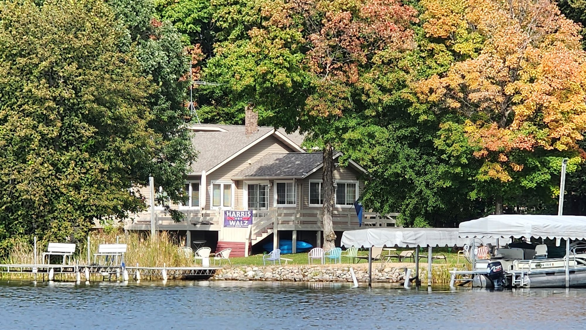 Orange, red, lime green colors envelope a home on a lake. A Harris Walz banner affixed to the deck surrounding the home. Boats in foreground.