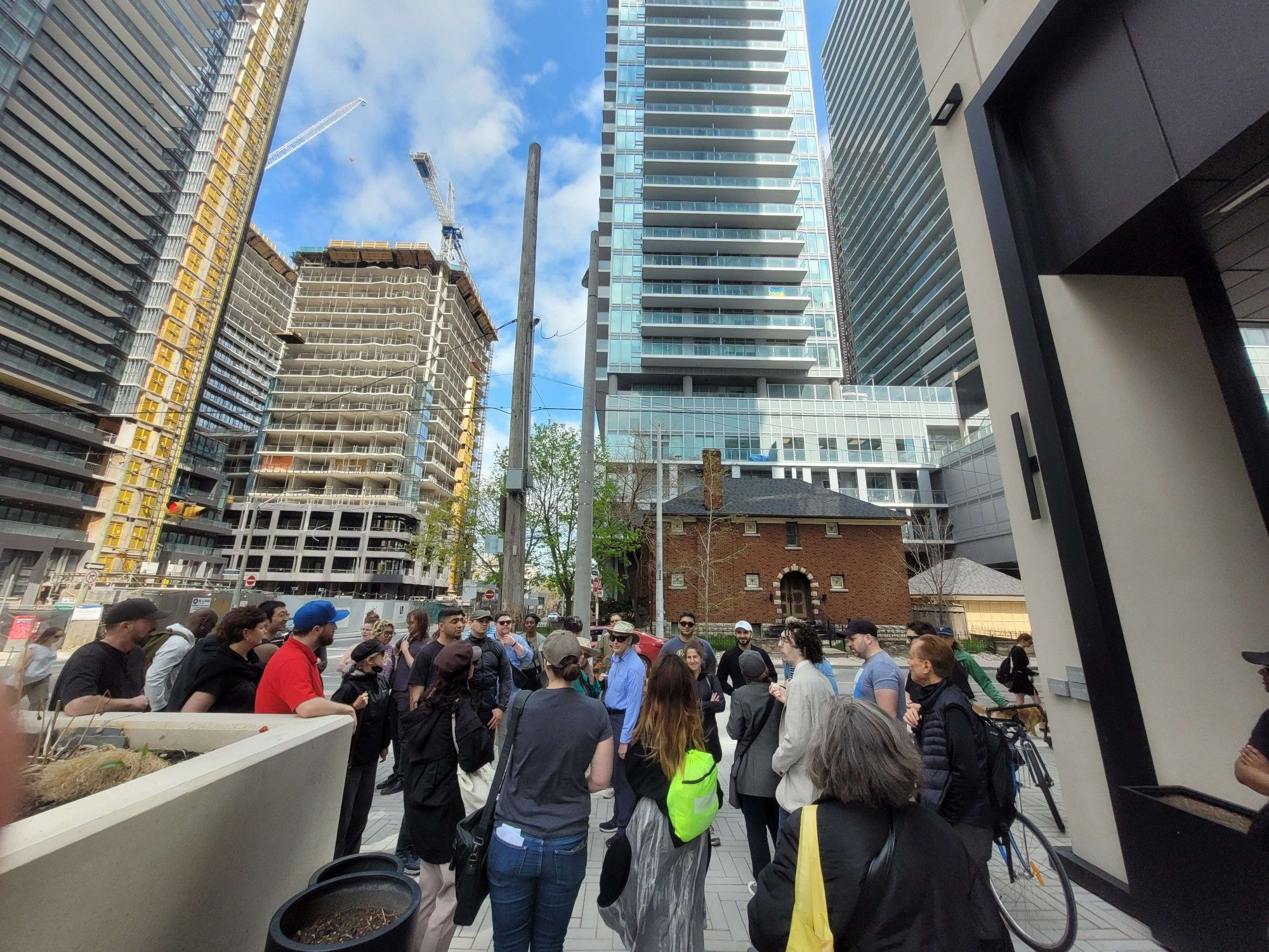 An image - distorted by a lens to capture more of several tall high-rises in the background - of a group of about 40 people beside a cafe. Across the street from them, a red-brick, two-storey house sits in front of a high-rise that is cut off in the image but appears to be about 30 storeys. Kitty corner, there are several cranes for other high-rises under construction.