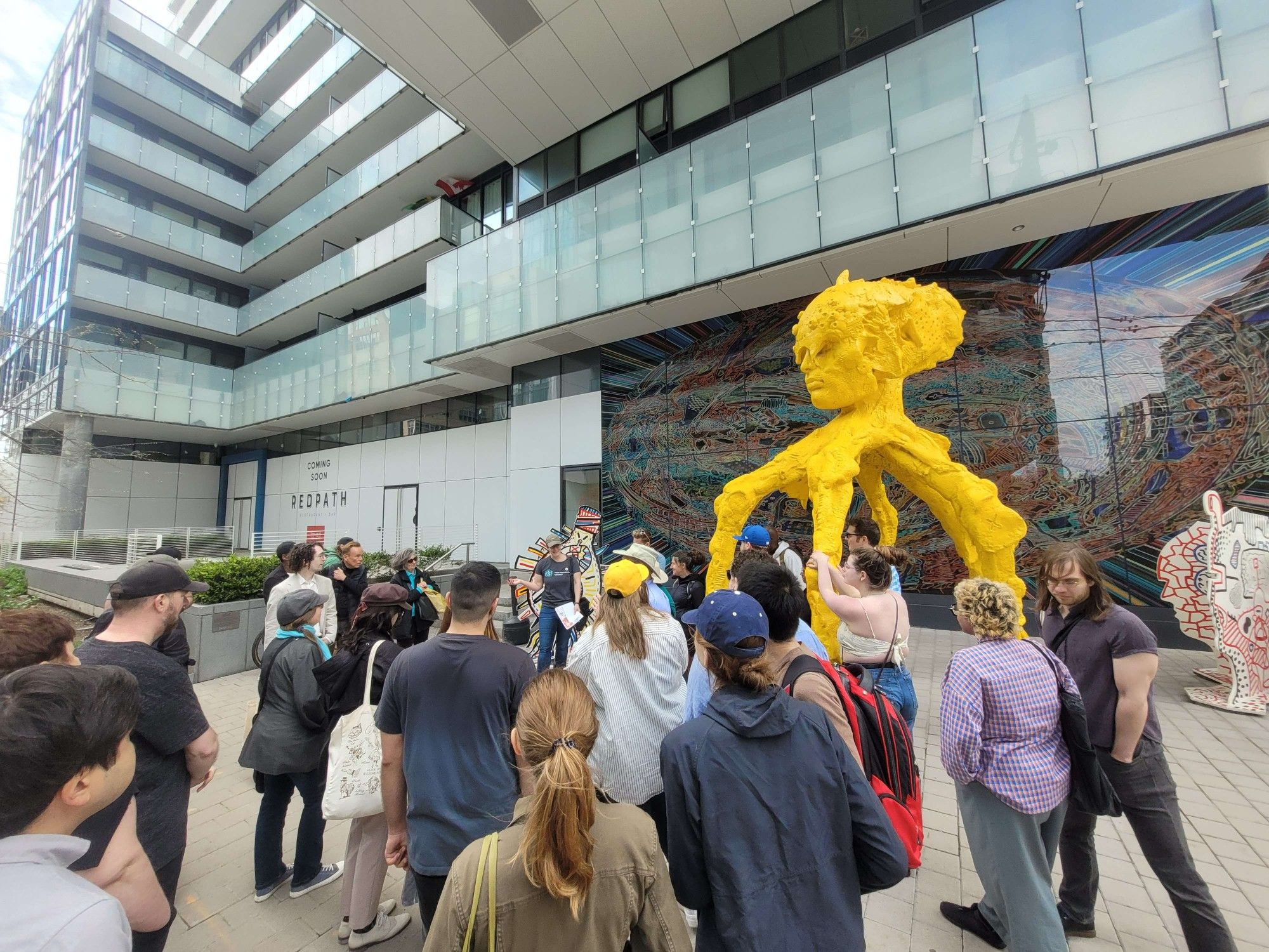 A group of people stand in the shade of a high-rise building, with the photo cutting off at about the tenth floor. Beside the group, is a large yellow alien-like sculpture, with several smaller, odd sculptures around and a mural behind. This is STARGATE by Blue Republic.