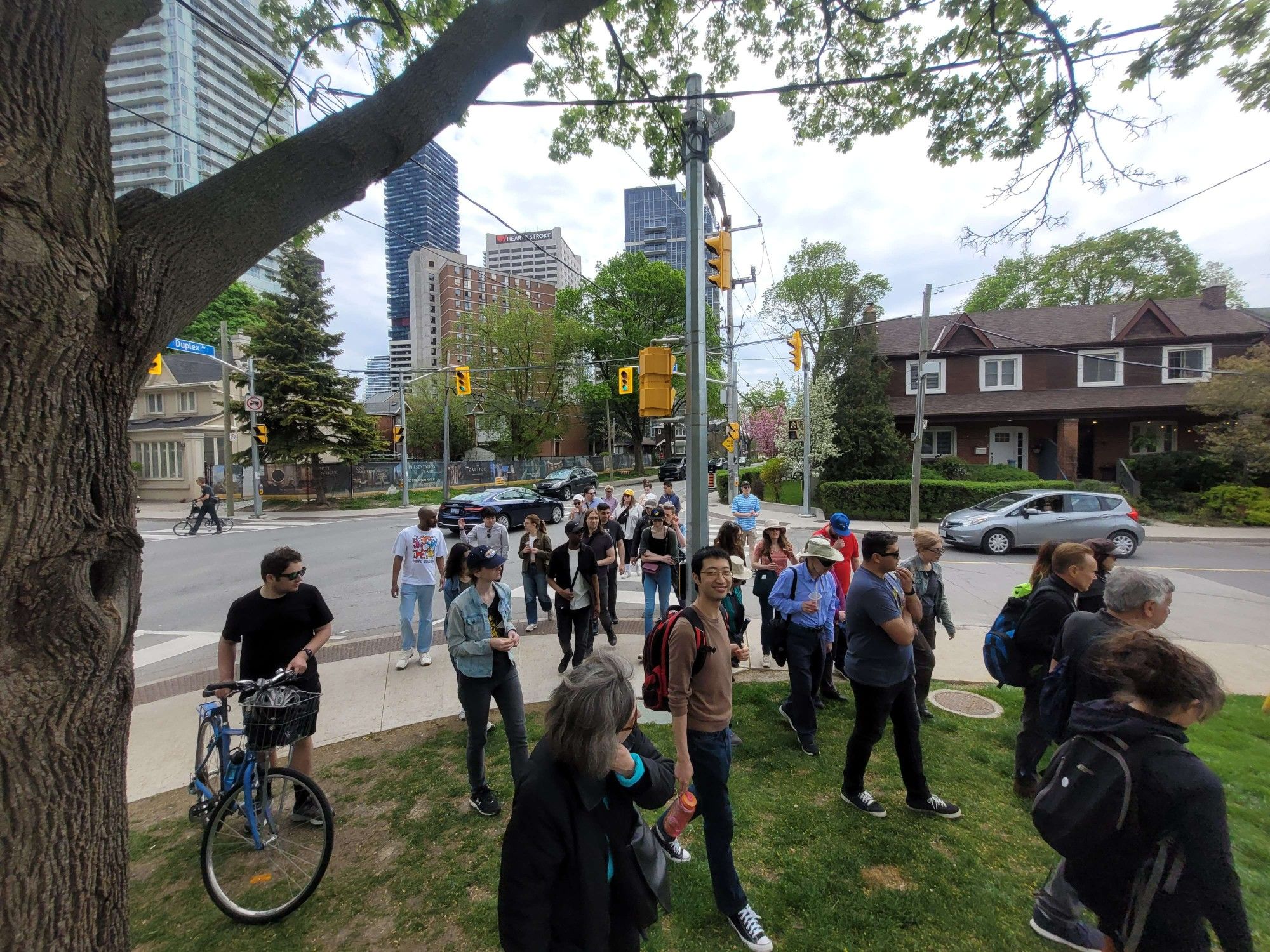 A group of people, including cyclists crosses at a light, up onto a grassy area. Directly behind them are several low-rise homes. In the distance, several high-rises can be seen.