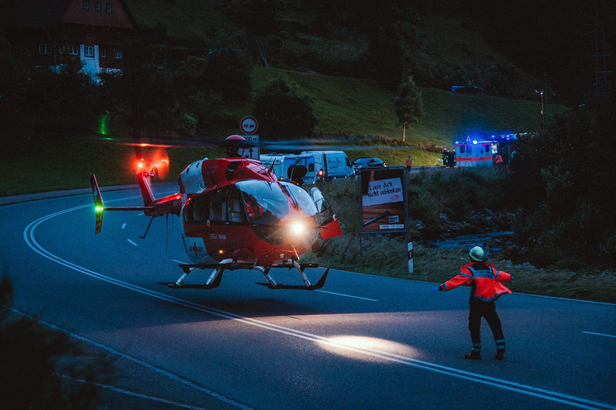 Bild eines landenden Rettungshubschraubers, ein Rettungsassistent weist den Hubschrauber ein. Es ist Dämmerung, im Hintergrund sieht man mehrere Rettungsfahrzeuge und eine Unfallstelle.