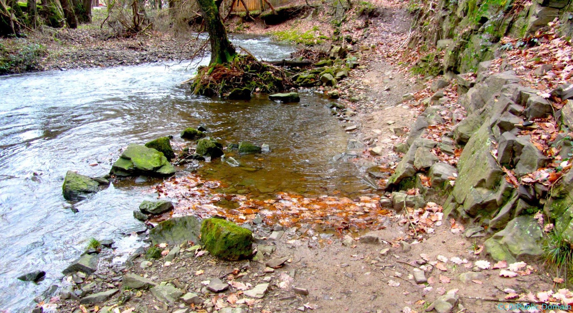 Links im Bild befindet sich der Vichtbach, rechts in unbefestigter Wanderweg, der vom Vichtbach überschwemmt wird. Rechts davon Felsen, etwa in der Mitte steht ein Baum im Wasser. Im oberes Bereich des Bildes biegt der Bach nach links ab.