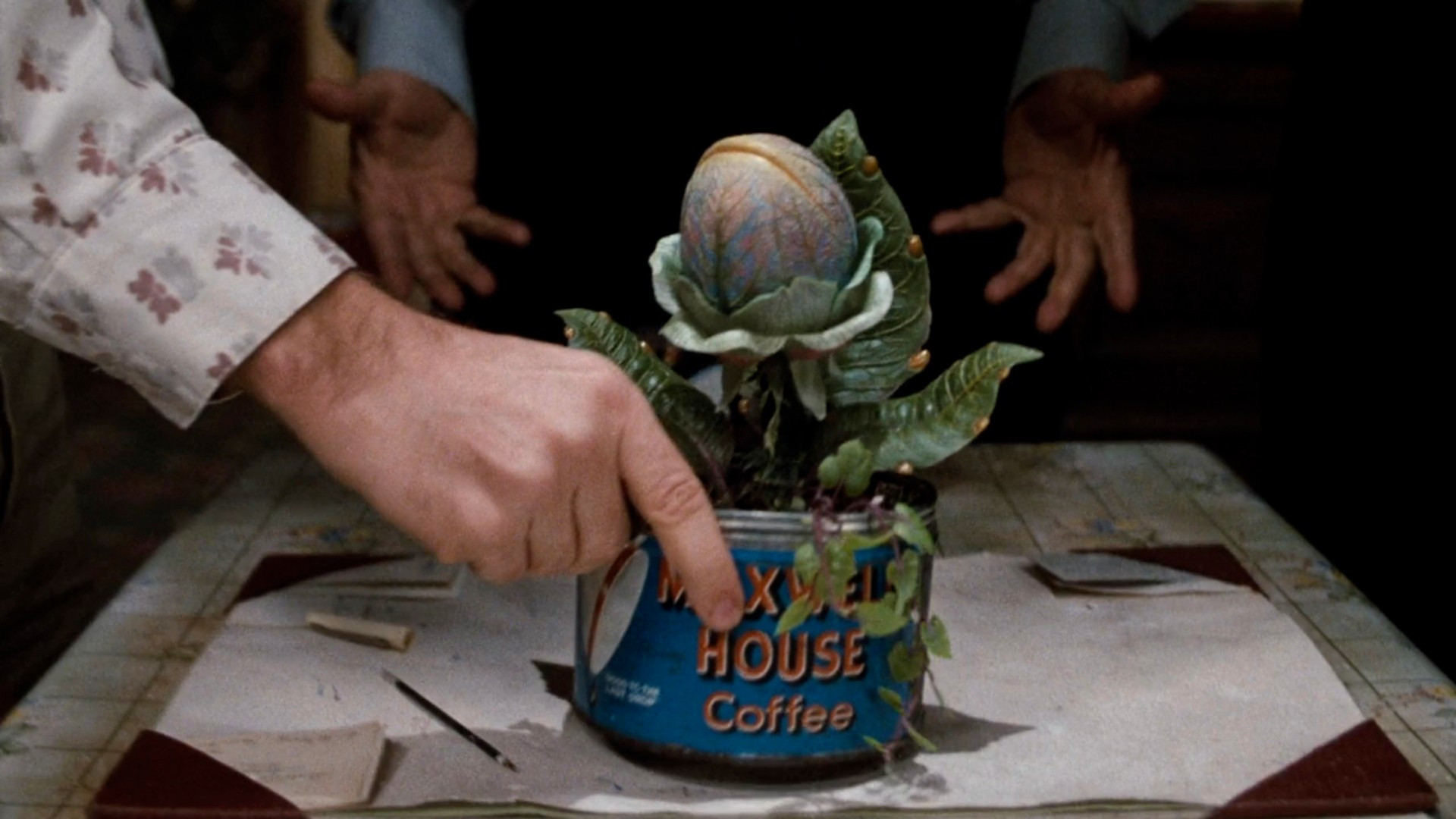 A hand reaches over to pick up a Maxwell House coffee can, which holds a strange, bulbous alien plant