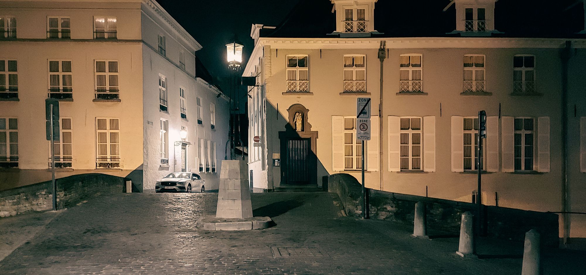 Nightshot of a cobble-stoned bridge showing a street lantern in the middle and back, buildings on either side, and a parked car.  