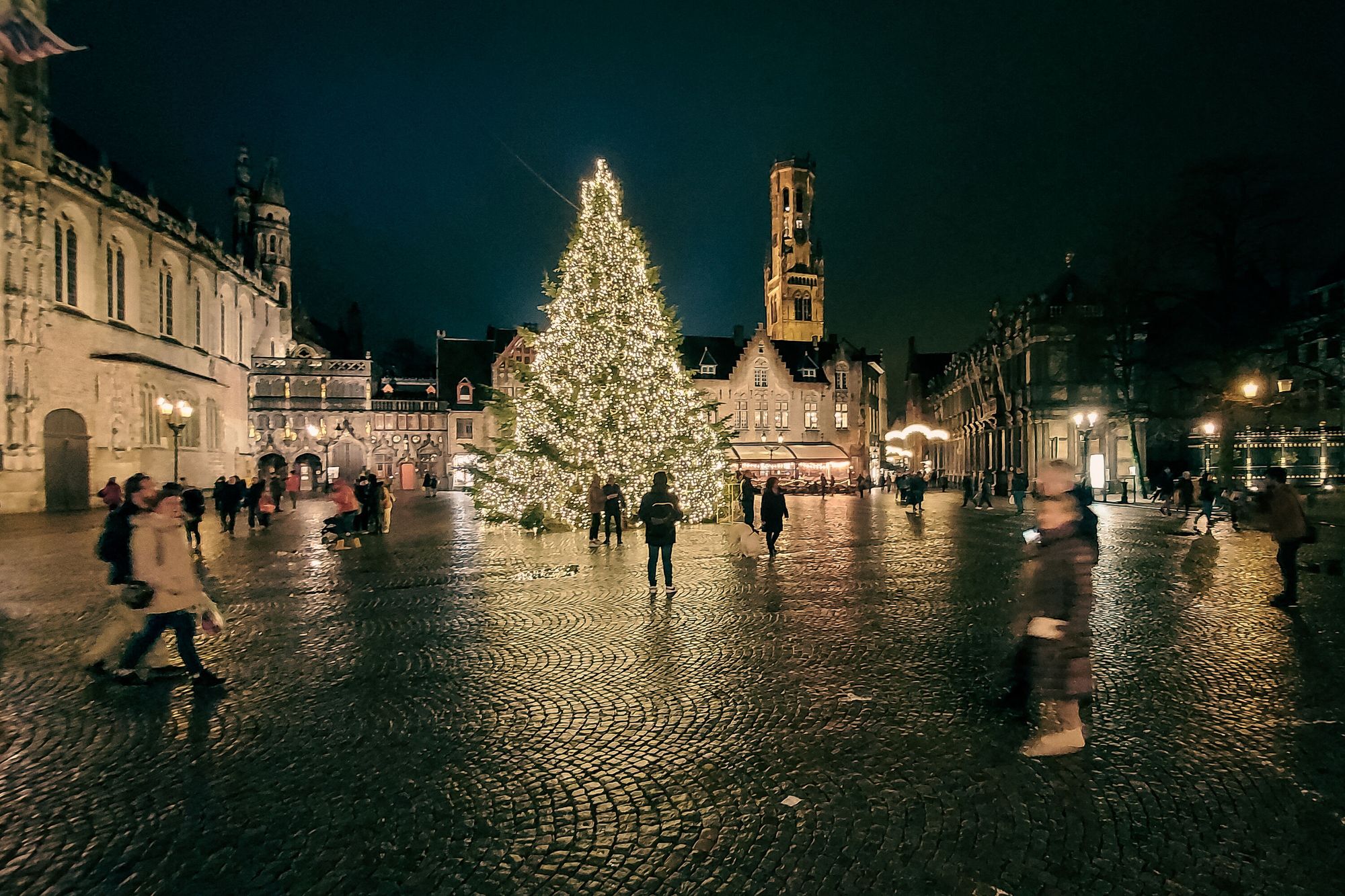 Nightshot of a large cobbled square with medieval buildings and a brightly-lit Christas tree.  The Belfort tower is visible in the background.