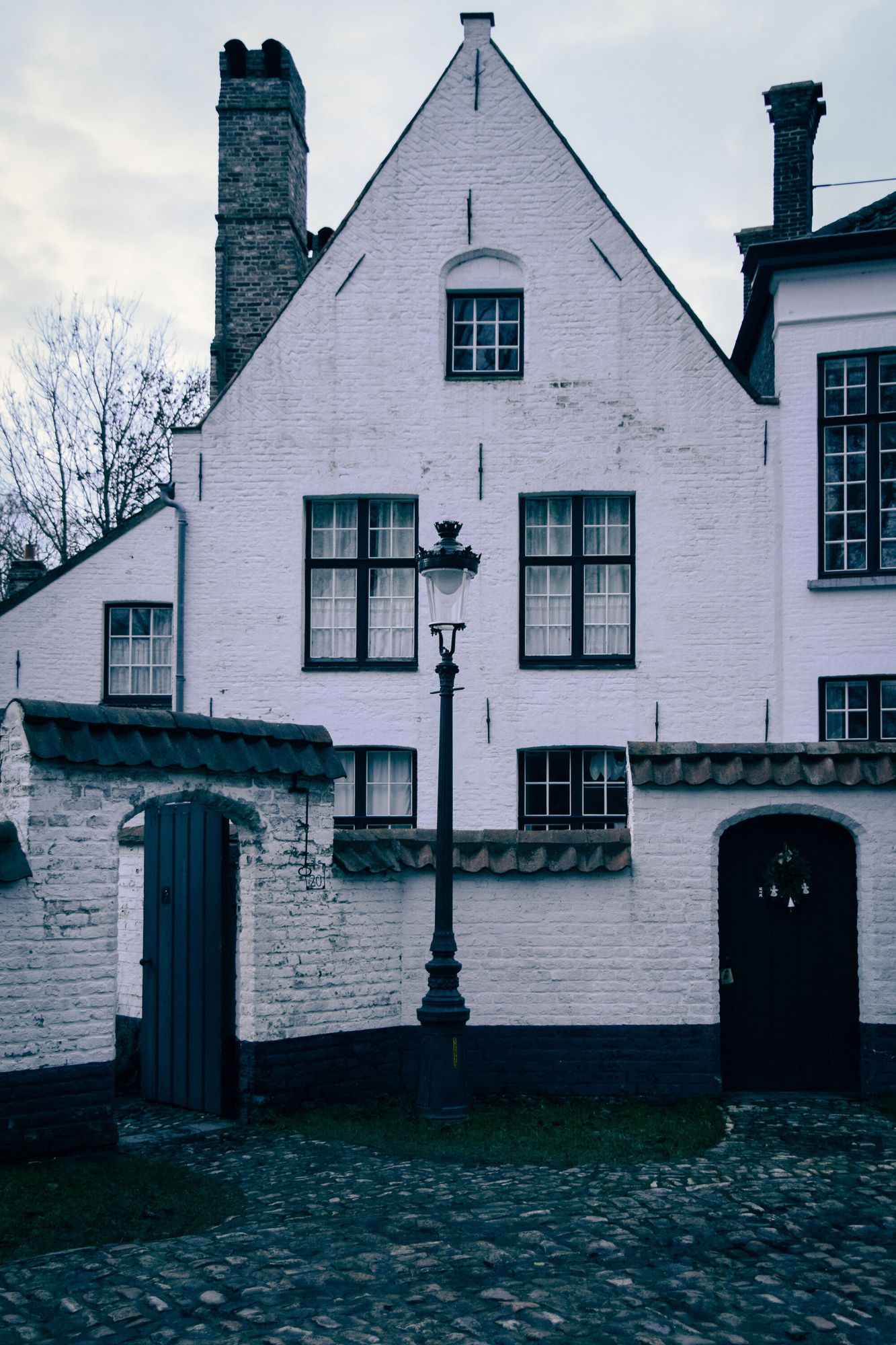 An old brick house painted white behind two gates (one open), a lamppost on a cobbled street at dusk.