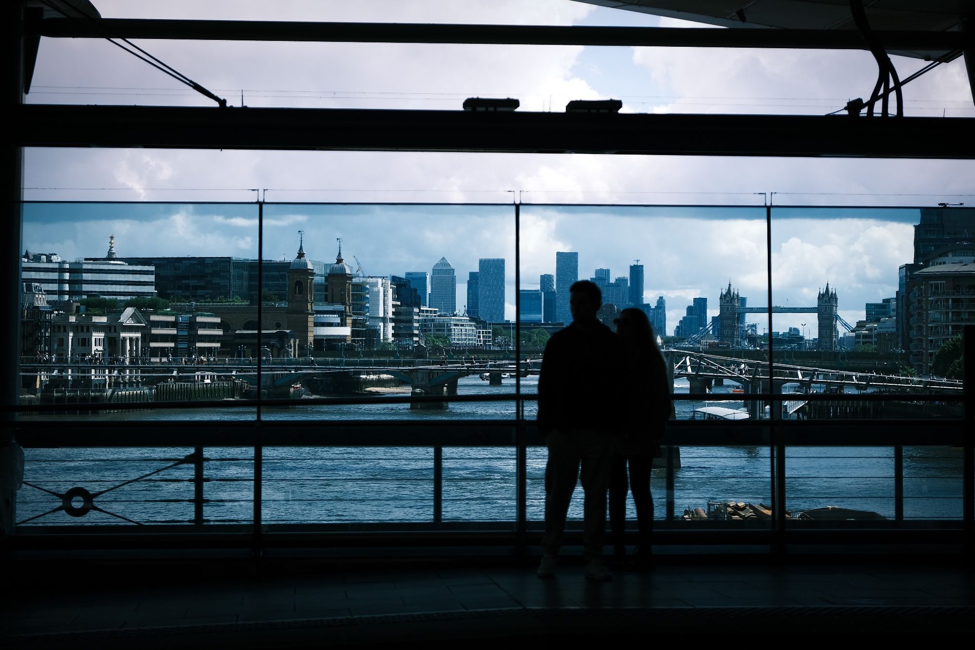 A cinematic, largely blue-toned colour photo of a silhouetted couple looking out over the River Thames from London's Blackfriars Station.