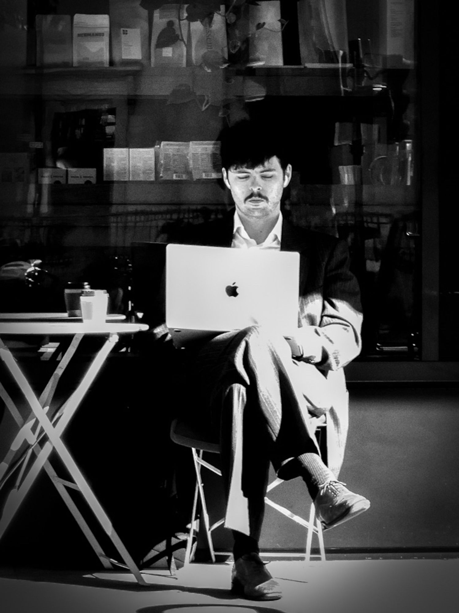 A high contrast black and white photo of a man sitting at a table outside a cafe using a MacBook on his lap.