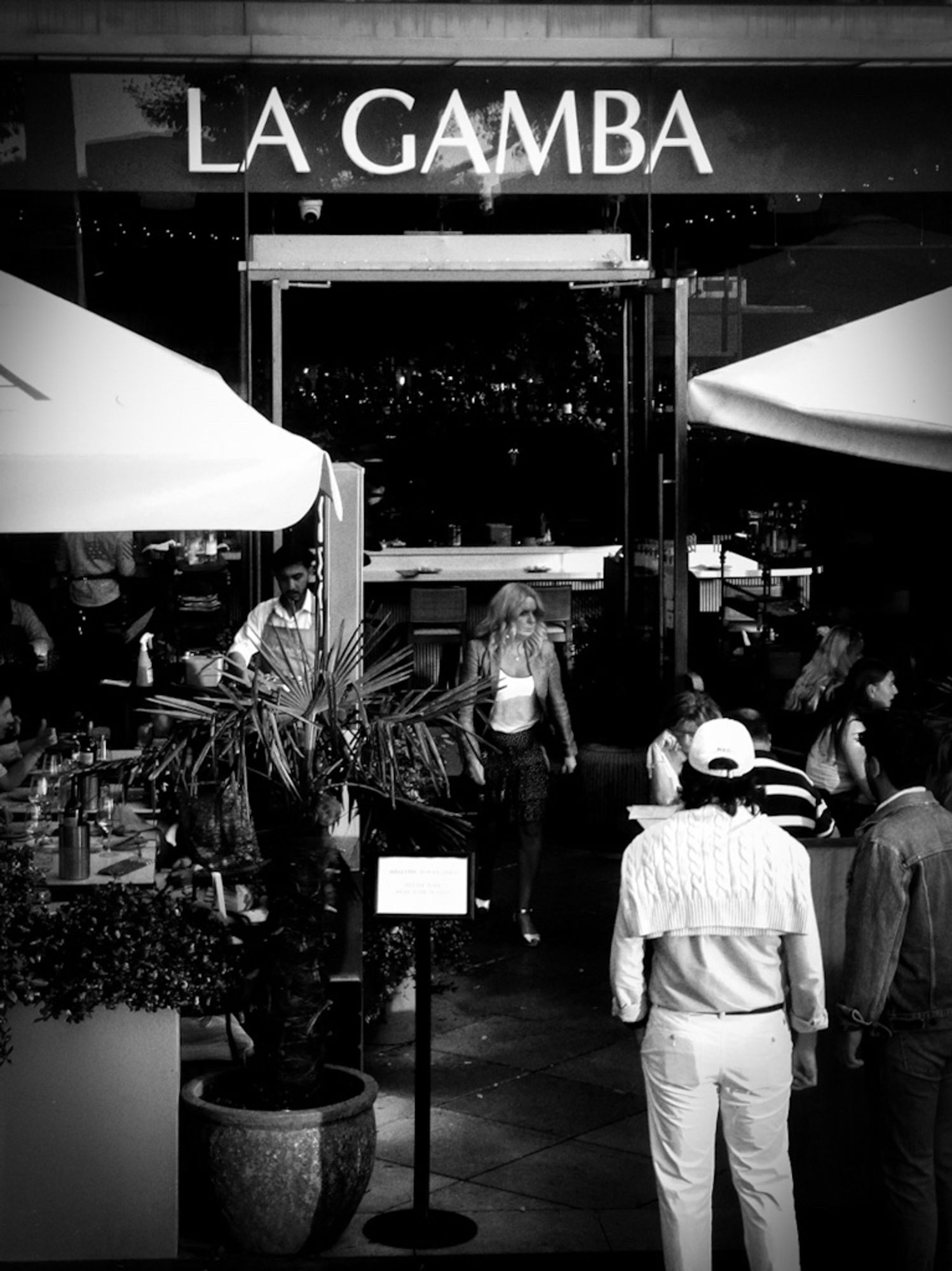 A high contrast black and white photo of a woman coming out of a seafood restaurant at London's South Bank, called La Gamba, which is Spanish for The Prawn, while others outside wait to go in.