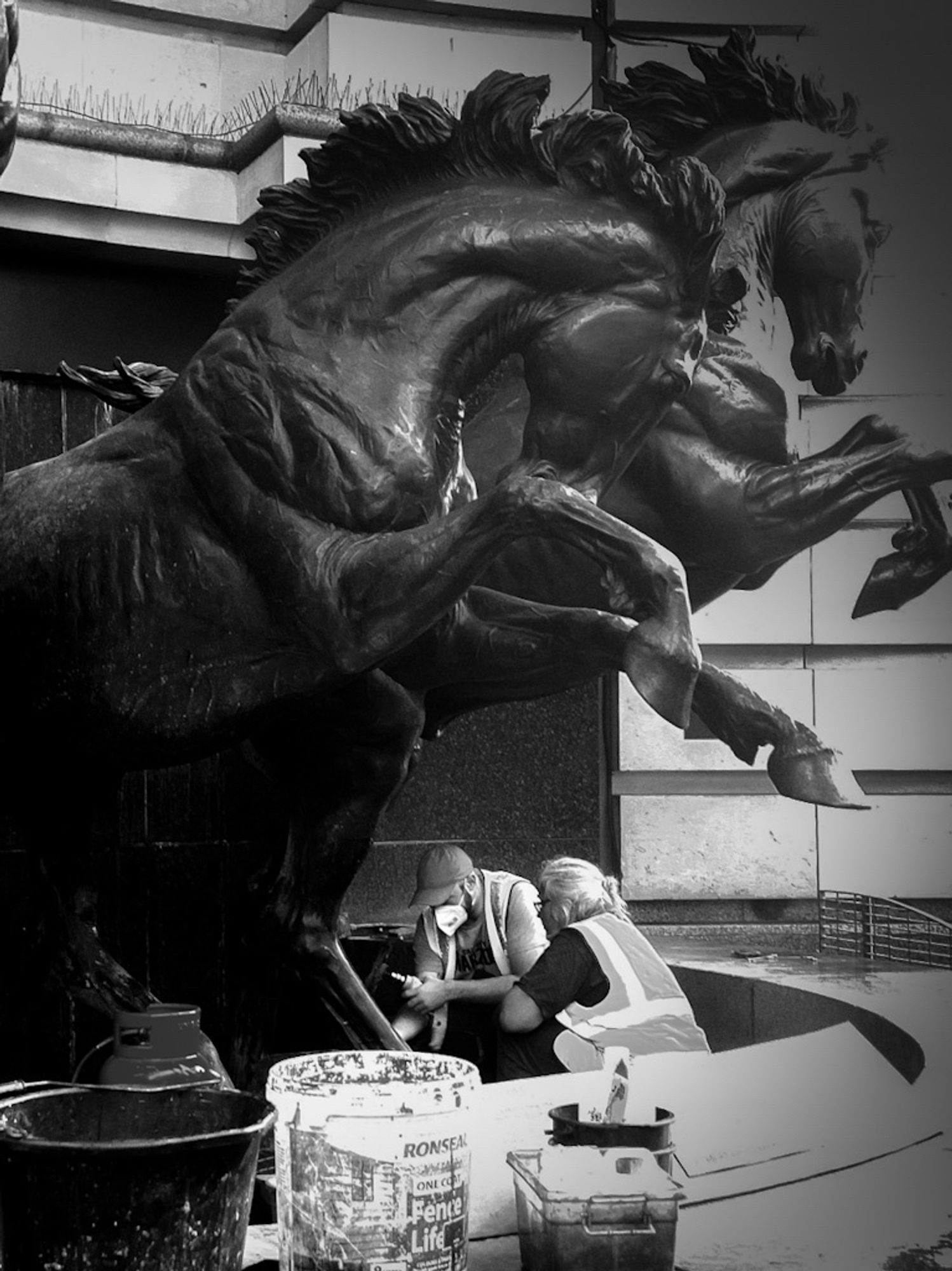 A high contrast black and white photo of two people repairing the feet of a pair of horse statues at London's Piccadilly Circus.