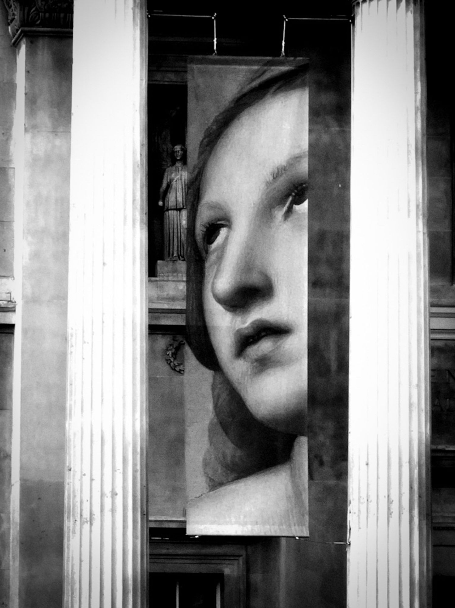 A high contrast black and white photo of a huge poster featuring a female face outside the National Gallery in London, with a much smaller statue of a woman to the left.