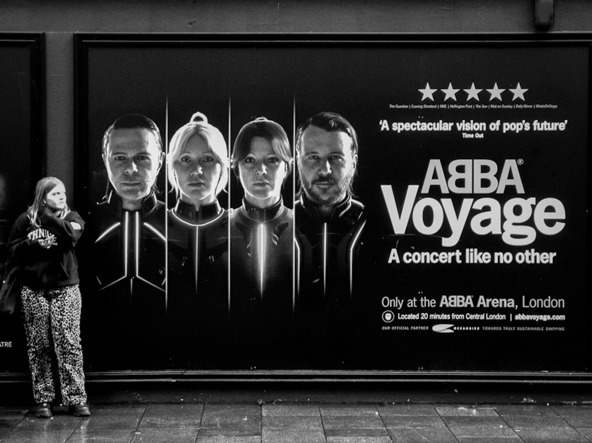 A high contrast black and white photo of a young woman standing next to a poster advertising the ABBA Voyage concert.