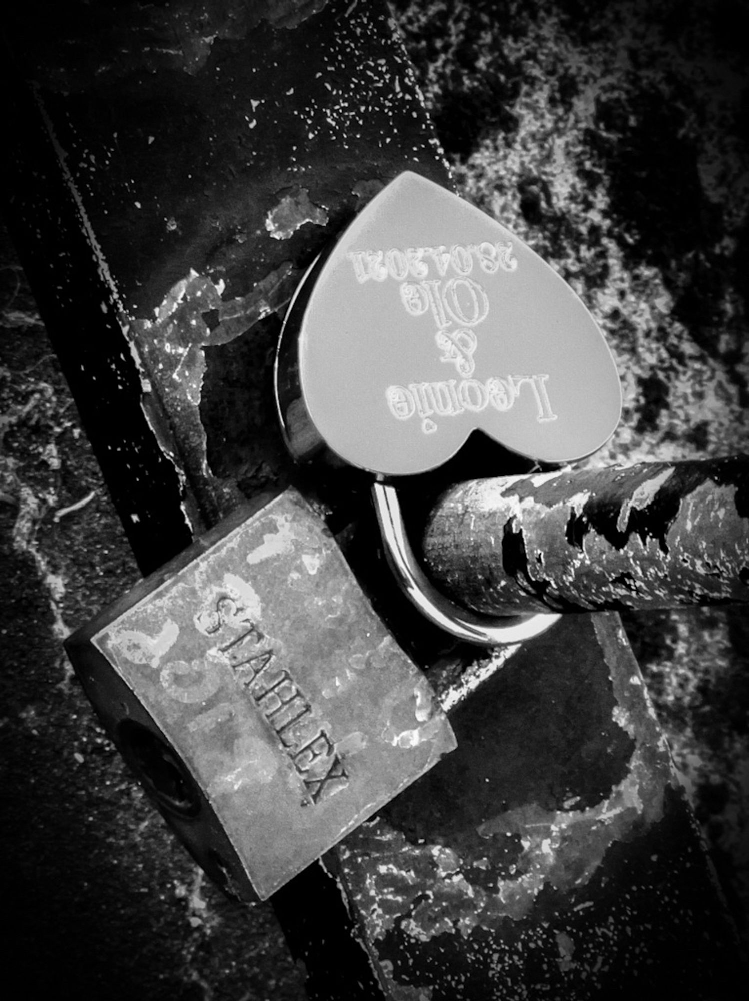 A high contrast black and white photo of two locks on a railing on the South Bank by the River Thames in London, one in the shape of a heart bearing the names of two people, the other a bicycle lock with the manufacturer's name, Stahlex.