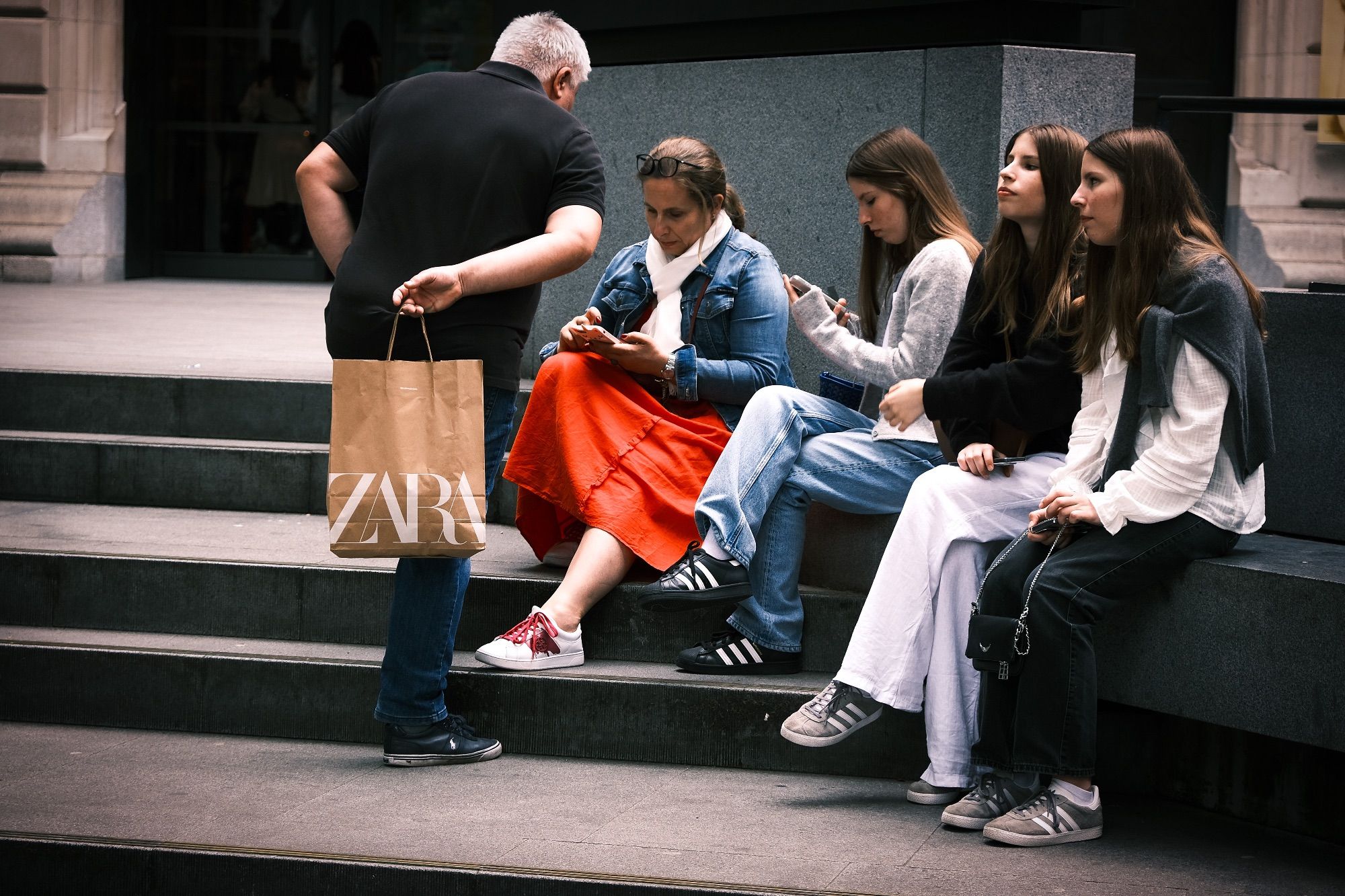 A colour photo of a family, consisting of middle aged mother and father with their three almost identical daughters aged somewhere around 16 years old, sitting on a stone bench outside London's National Portrait Gallery. The mother and one of the girls are looking at their phones, the father is standing, leaning down towards the mother as If to get her attention, and the two other girls are looking outwards at nothing in particular.