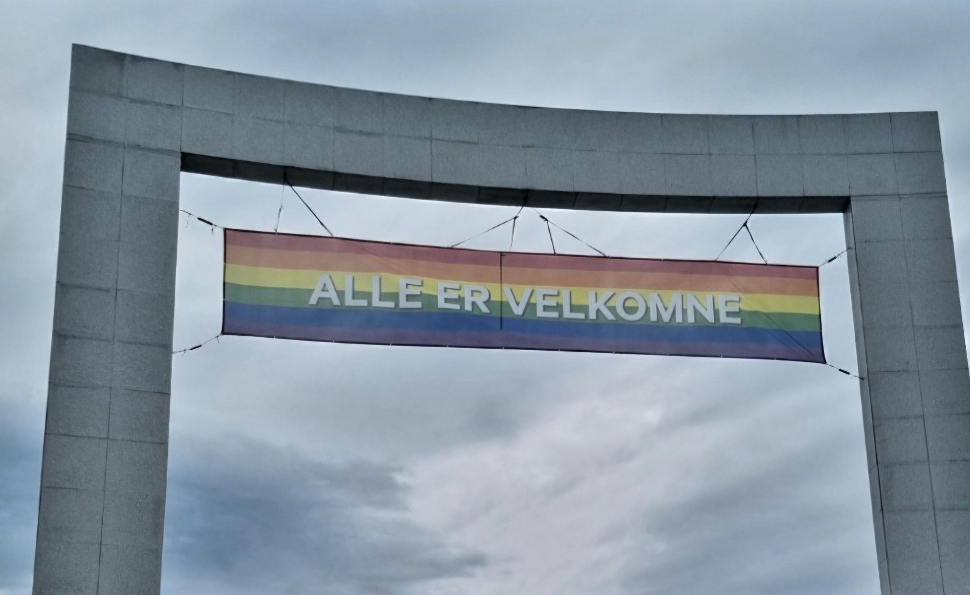 Ausschnitt eines Torbogens mit einem Regenbogenschild mit der Aufschrift "ALLE ER VELKOMNE" vor bewölktem Himmel.