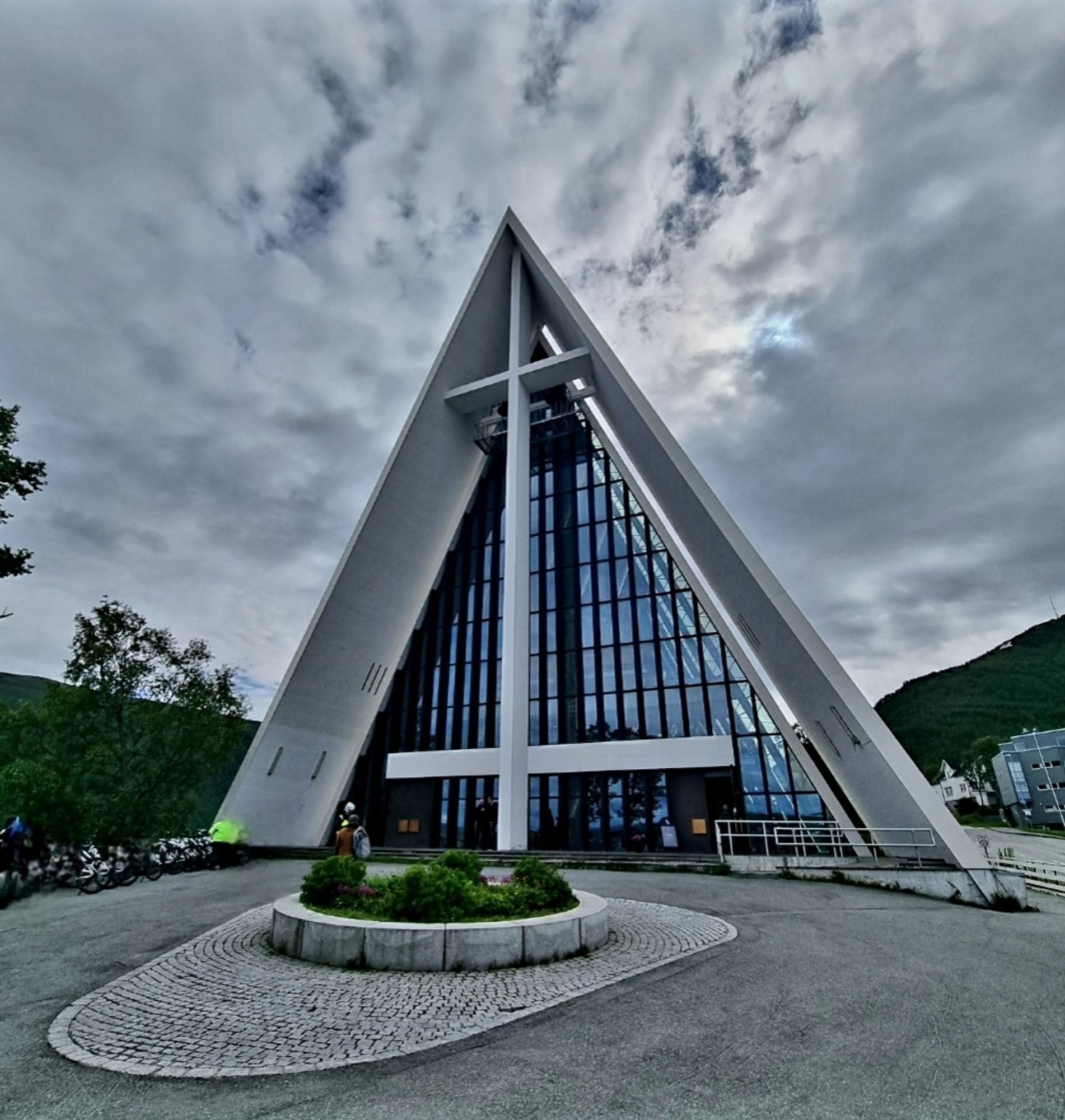 Dreiecksförmige Vorderansicht einer Kathedrale, die Front besteht aus rechteckigen Glasfenstern mit weißer Betonumrandung. Davor Asphalt und ein kreisförmiges Blumenbeet. Der Himmel ist bewölkt.