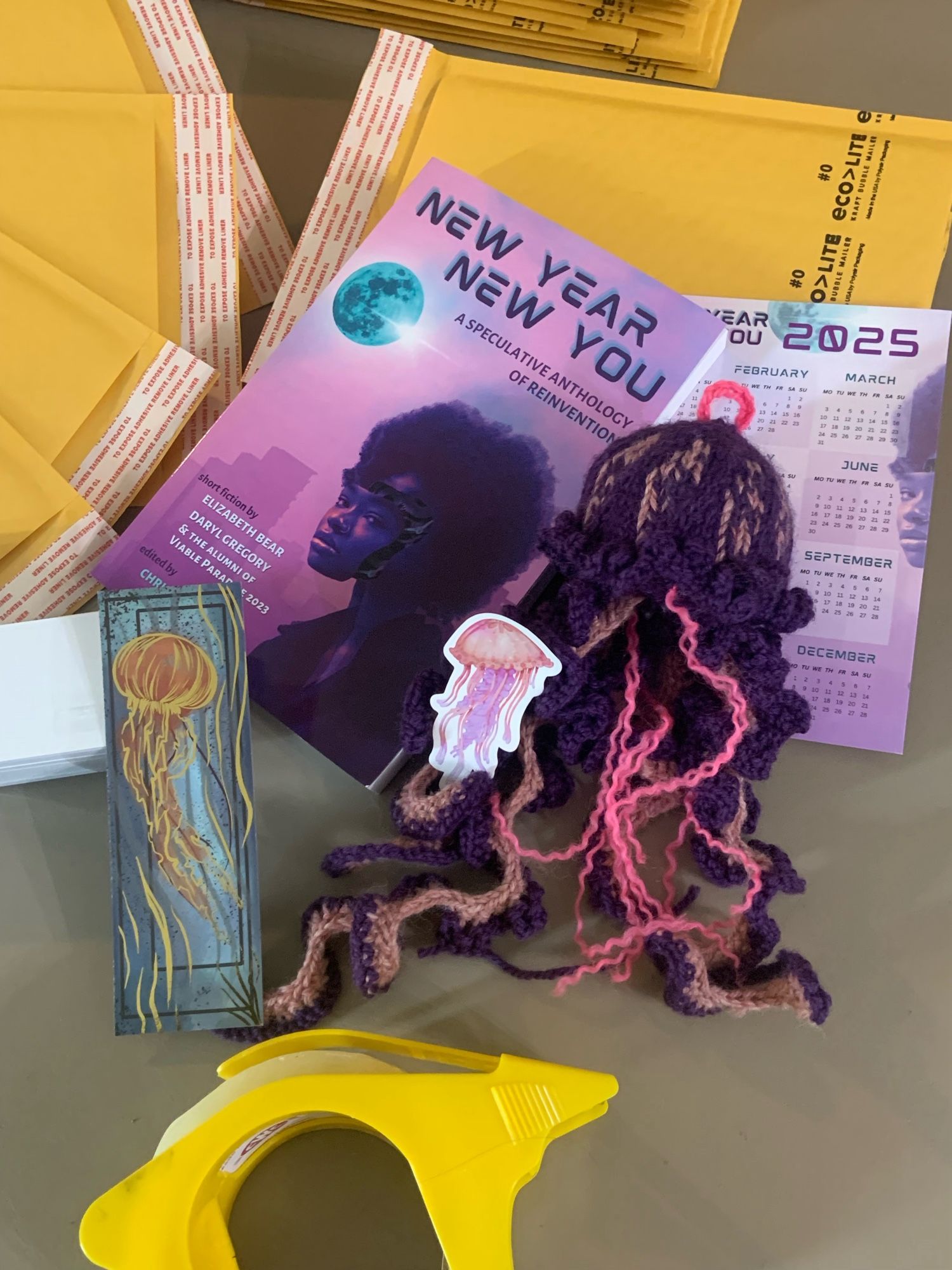 A spread of kickstarter rewards. A purple anthology titled: New Year, New You. A purple knit jellyfish. A foil bookmark. A calendar postcard. A jellyfish sticker. 

They are laid against packing envelopes.