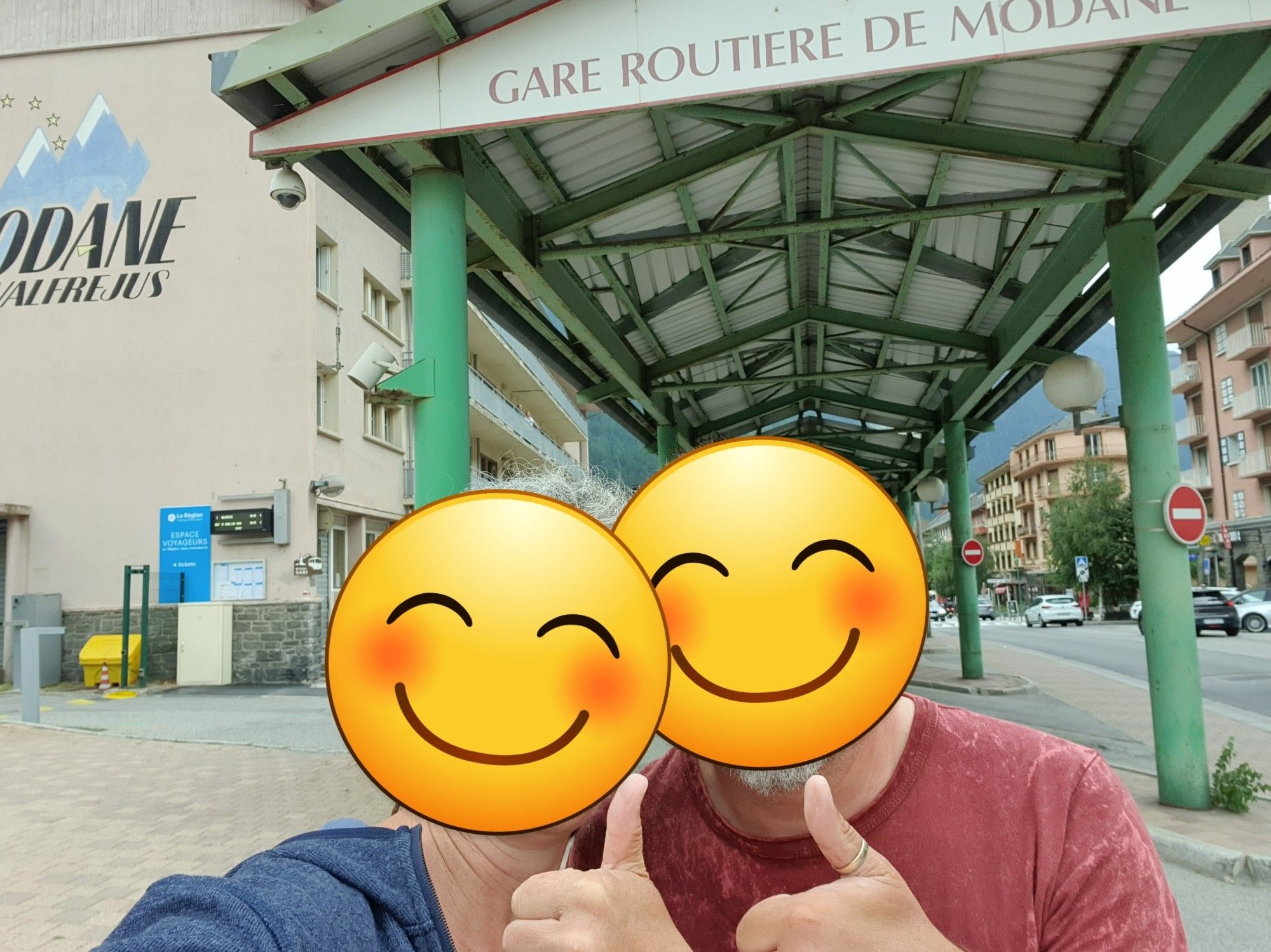 Die Userin und ihr Mann im Selfie-Porträt vor einem hohen metallenen Dach, auf dem "Gare Routière de Modane" steht. Links davon ein beiges mehrstöckiges Gebäude, rechts eine Straße.