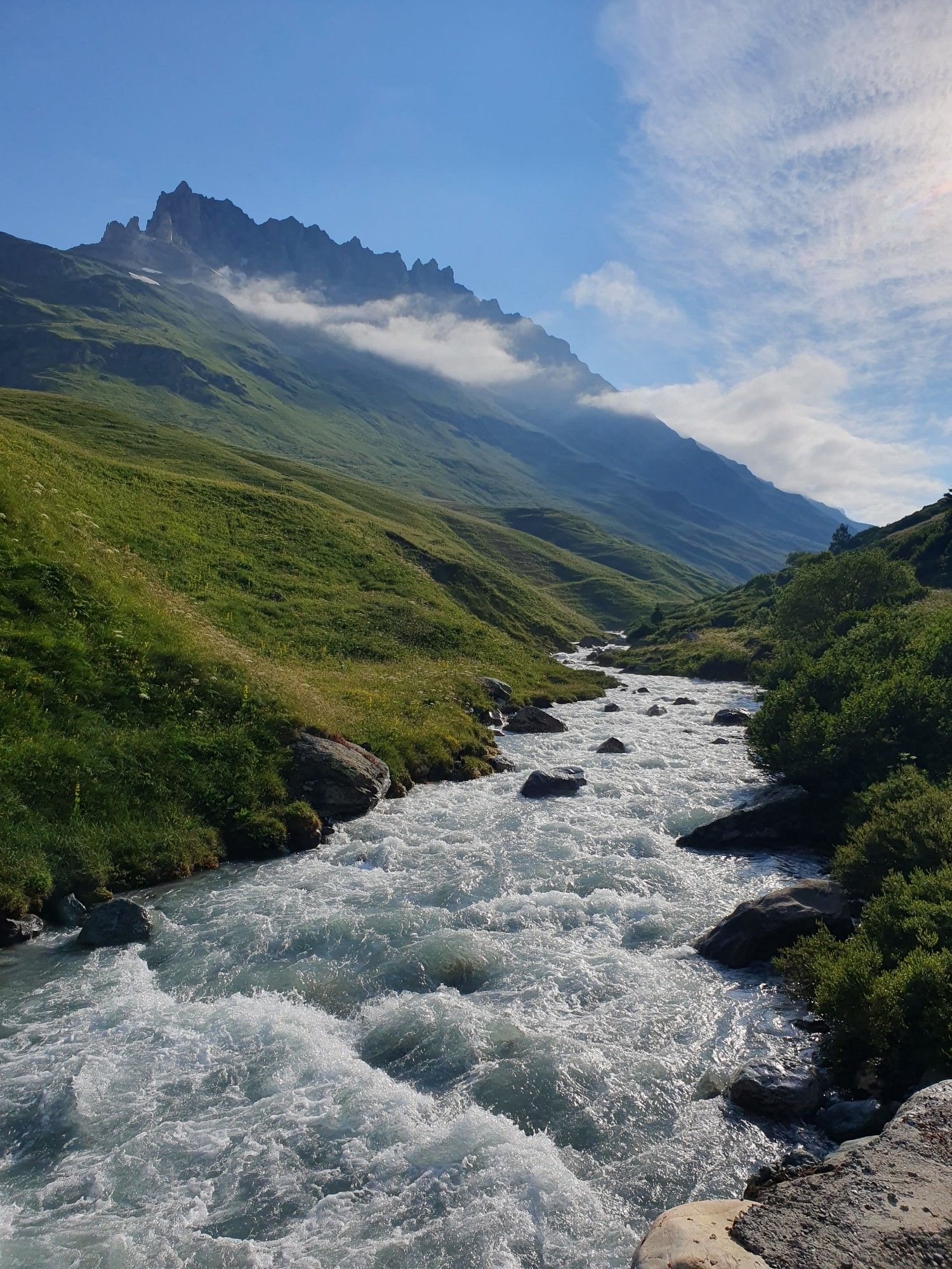 Im Vordergrund fließt ein breiter Alpenbach auf den Betrachtenden zu. Links und rechts grüne Hügel, im Hintergrund hohe felsige Berge, darüber blauer Himmel mit Wolkenschleiern.