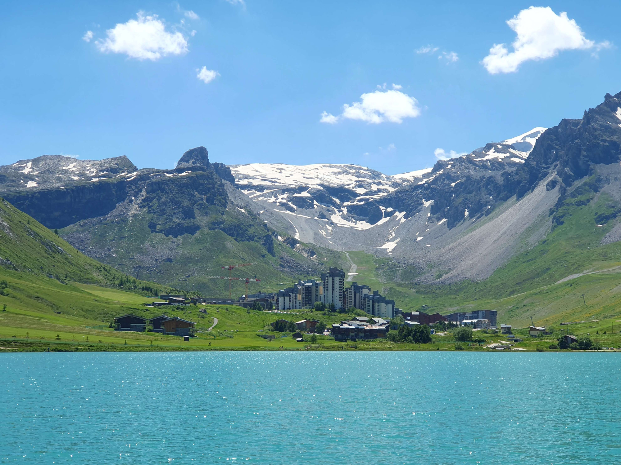 Türkisfarbener See vor Berglandschaft (unten Wiese, oben Felsen und Schnee) unter blauem Himmel mit drei weißen Wattewölkchen.

Auf der anderen Seeseite erkennt man eine Ballung großer Hotels.