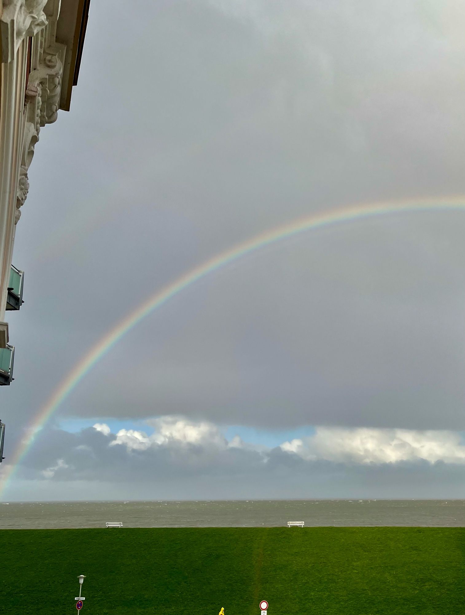 Der Abendhimmel zeigt einen Regenbogen, auf dem Deich sind zwei leere Bänke zu sehen 