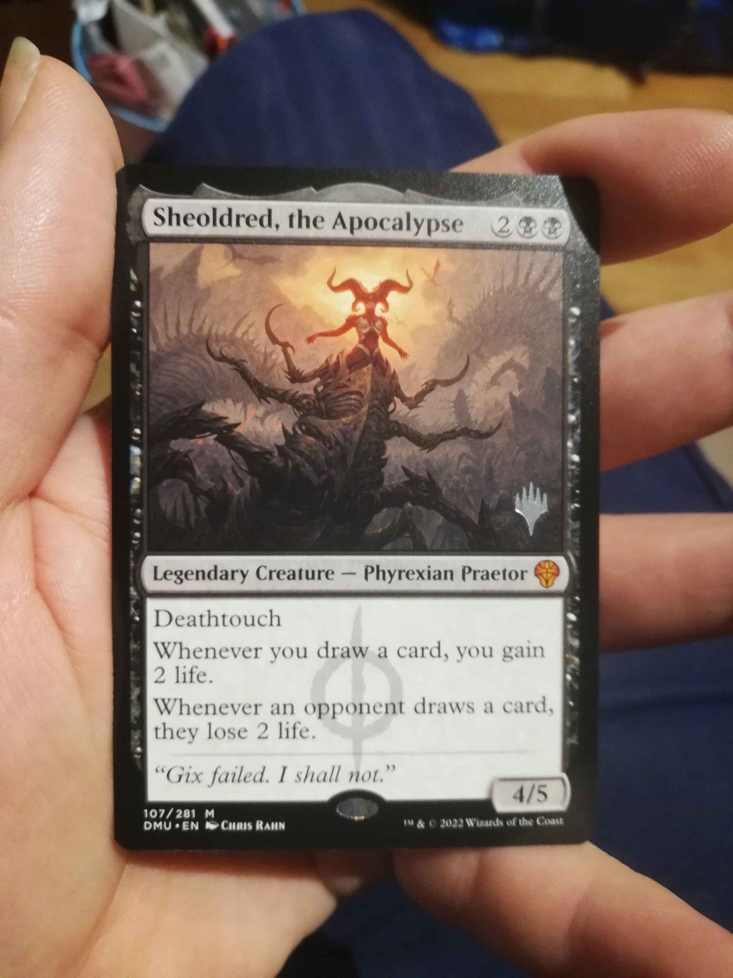 A picture of me holding the Magic: the Gathering card Sheoldred, the Apocalypse
It's 2BB for Legendary Creature Phyrexian Praetor that's a 4/5 with deathtouch and "Whenever you draw a card, you gain 2 life. Whenever an opponent draws a card, they lose 2 life."