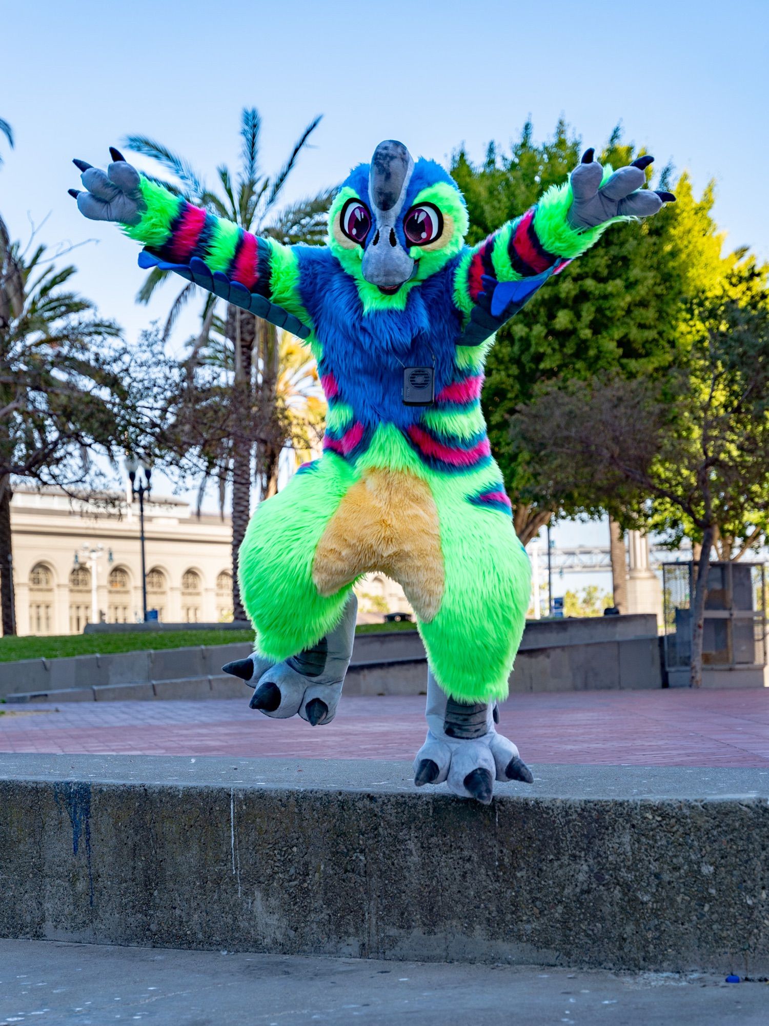 Blue and Green Citipati (an Oviraptorid Dinosaur) Fursuit jumping from a ledge with it’s arm spread wide as if it’s about to fly
