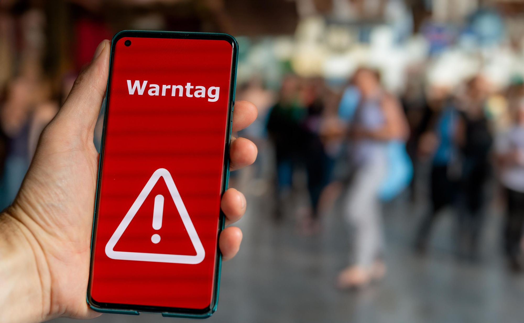 Eine Hand hält ein Mobiltelefon mit rotem Bildschirm auf dem „Warntag“ zu lesen ist sowie ein Dreieck mit einem Ausrufezeichen zu sehen ist. Im Hintergrund stehen Menschen. Quelle: Adobe.stock/Fokussiert