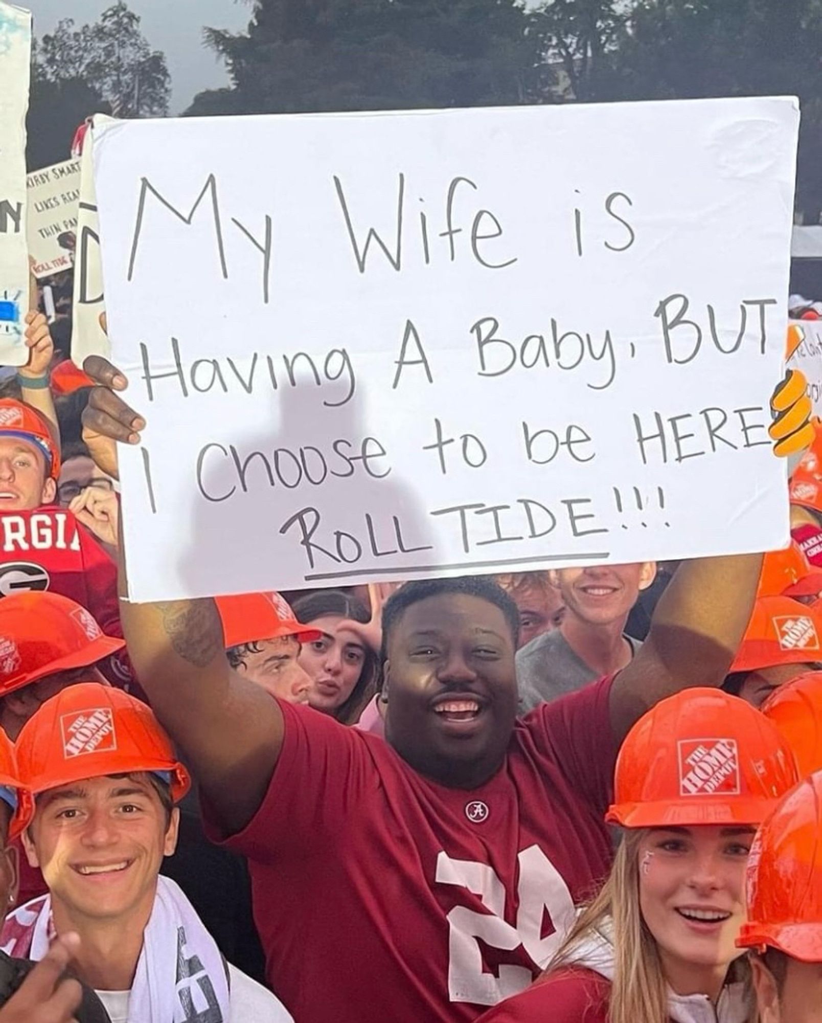 In a crowd of football fans, a man holds up a sign reading, “My Wife is Having a Baby, BUT I Choose to be HERE, ROLL TIDE!!!”