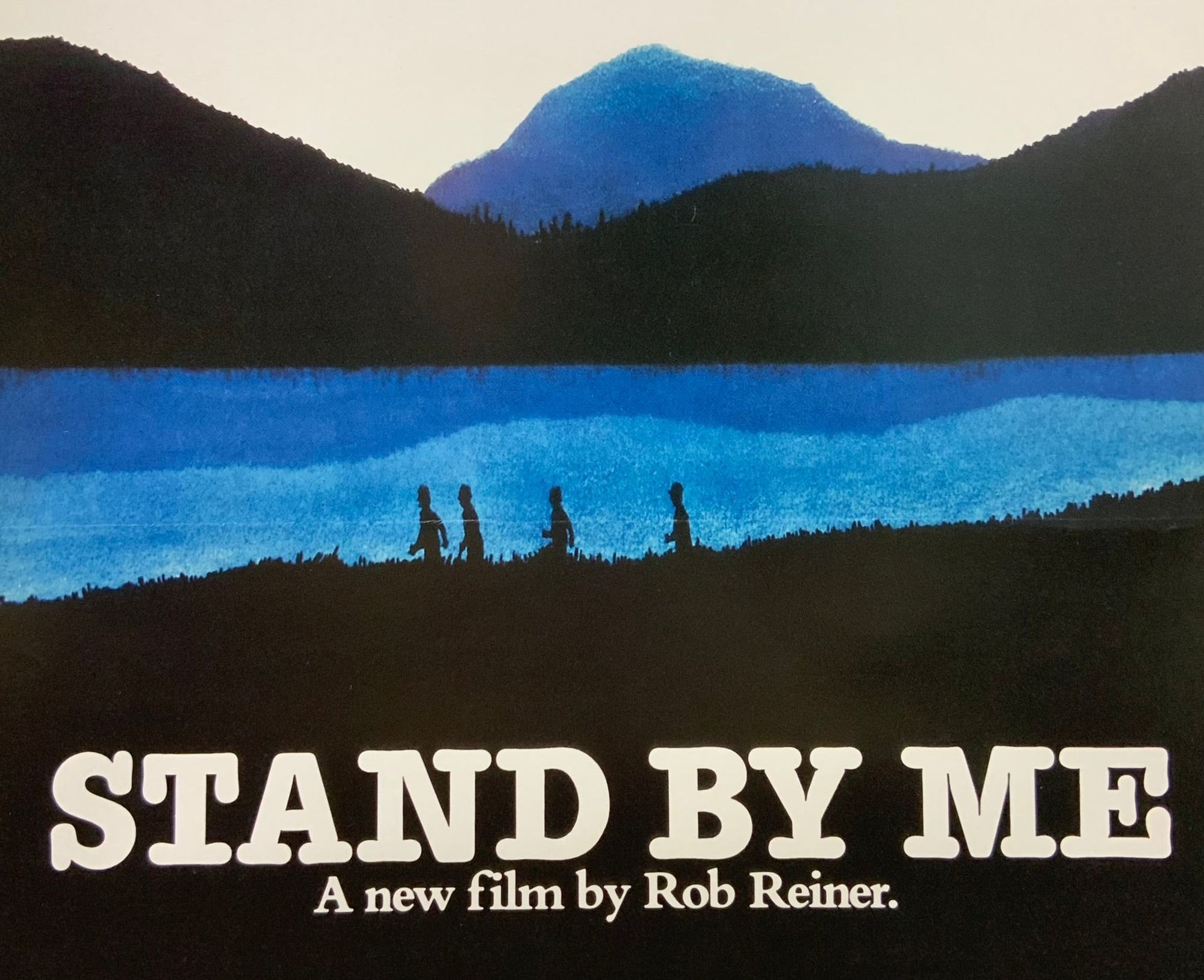 A blue mountain in the distance with rolling black hills in front of it. A huge blue lake with four boys' small silhouettes against it. Large black grassland in the foreground. Very big white text that says "Stand By Me" at the bottom of the poster. Below that smaller white text that says "A new film by Rob Reiner".