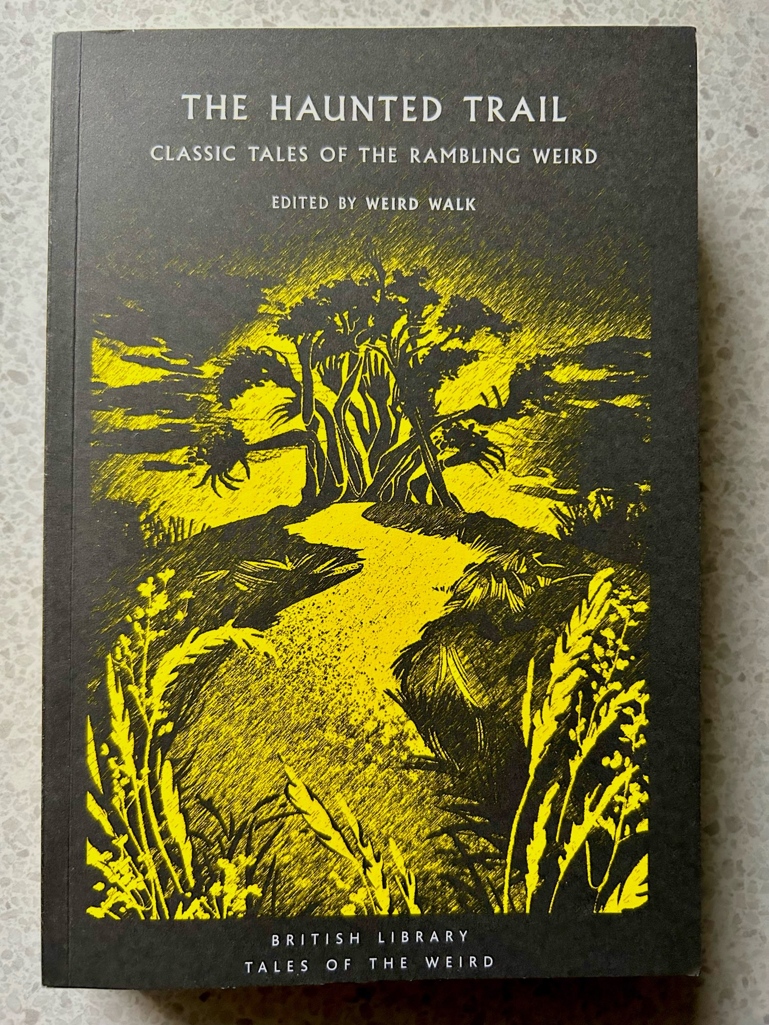 The front cover of a book of ghost stories called The Haunted Trail. Classic tales of the rambling weird. It features a yellow and black illustration of a path leading to a copse of bent trees.