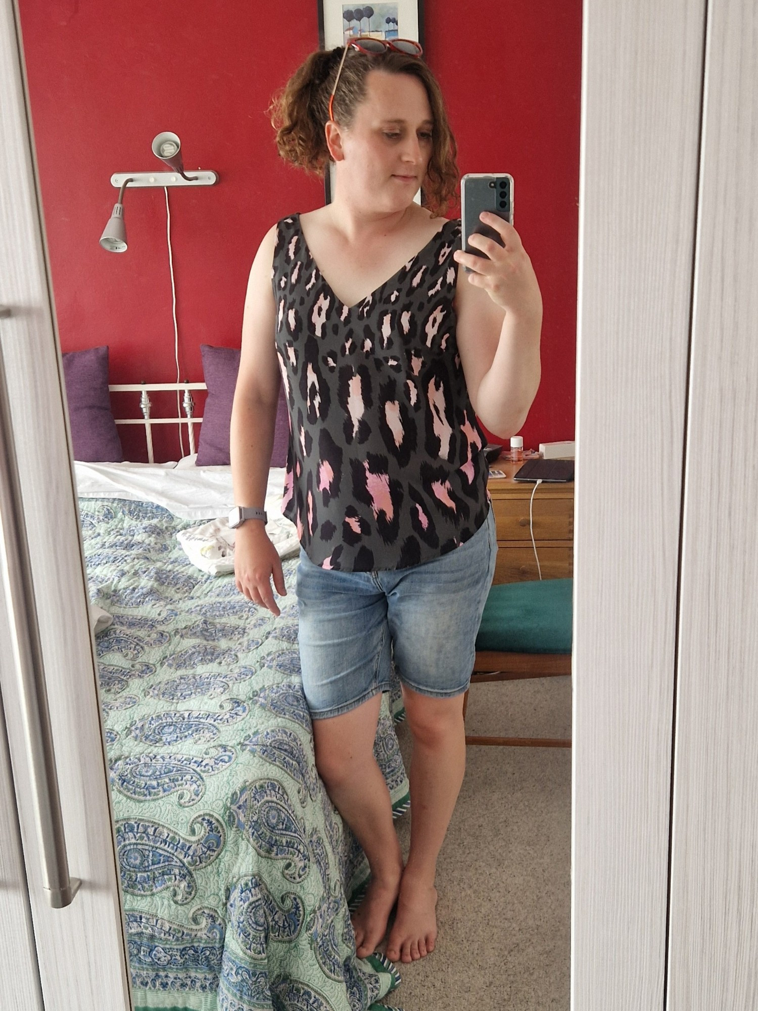 A full length picture of me taken in a mirror. I'm stood by a bed with a red wall behind me. I'm wearing denim shorts and an olive green animal print top. My hair is tied back except for a side fringe. I'm wearing red sunglasses on top of my head.