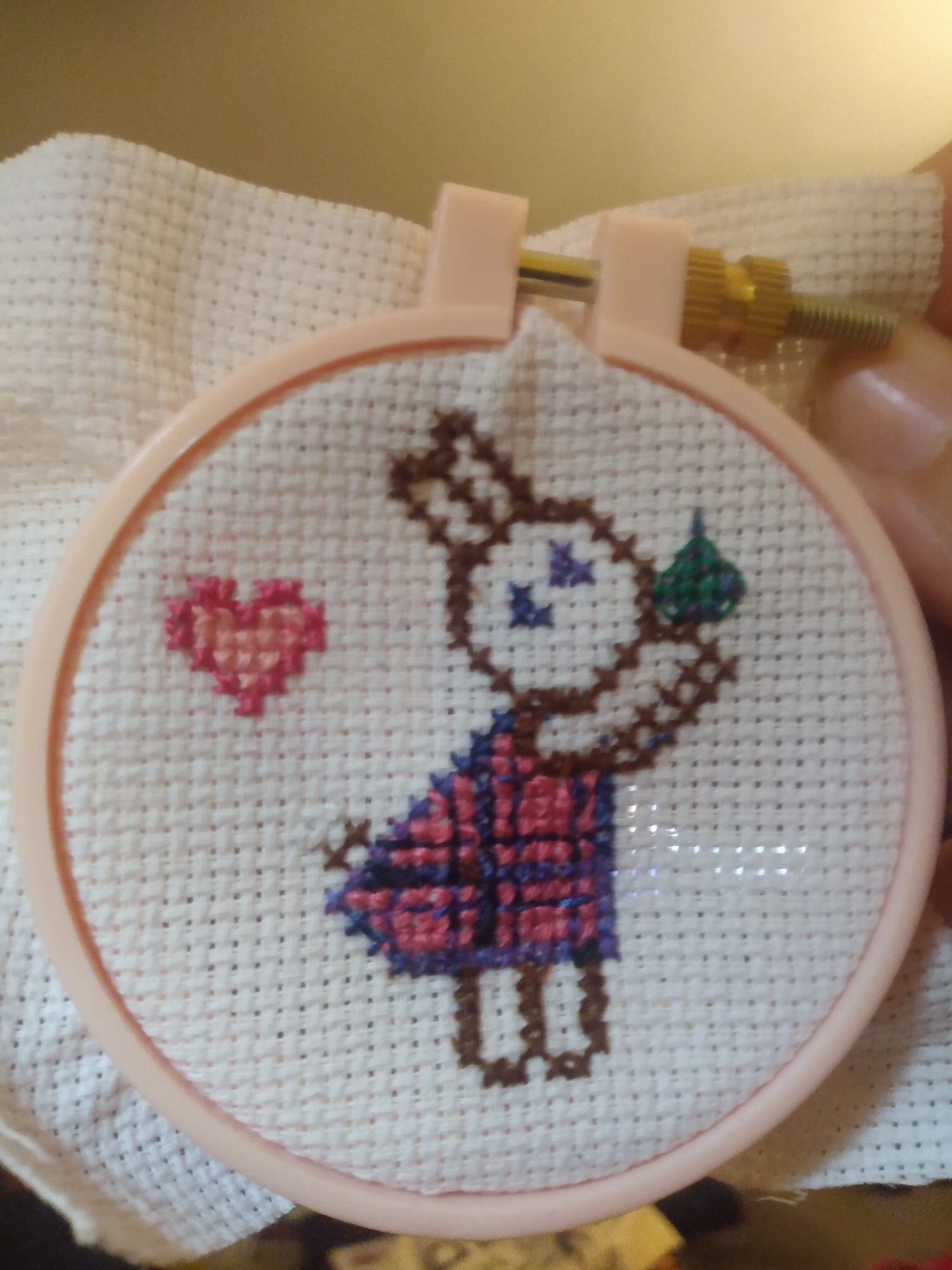 A cross stitch of a cartoon bunny in a plaid dress, holding up a paw paw fruit. A heart is in the left