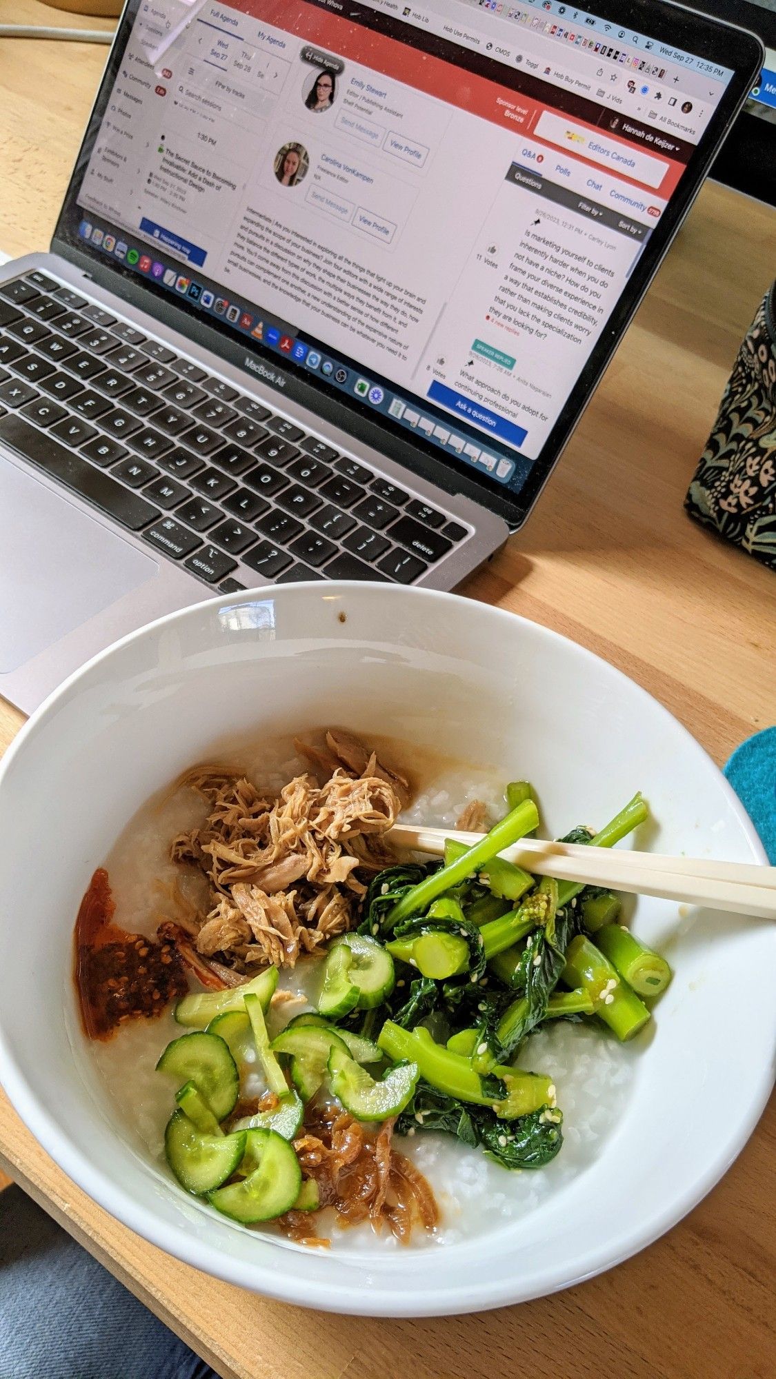 A big white bowl of congee, stewed chicken, cucumbers, pickled radishes, and greens with chopsticks sticking out of it on a wooden desk in front of an open laptop.