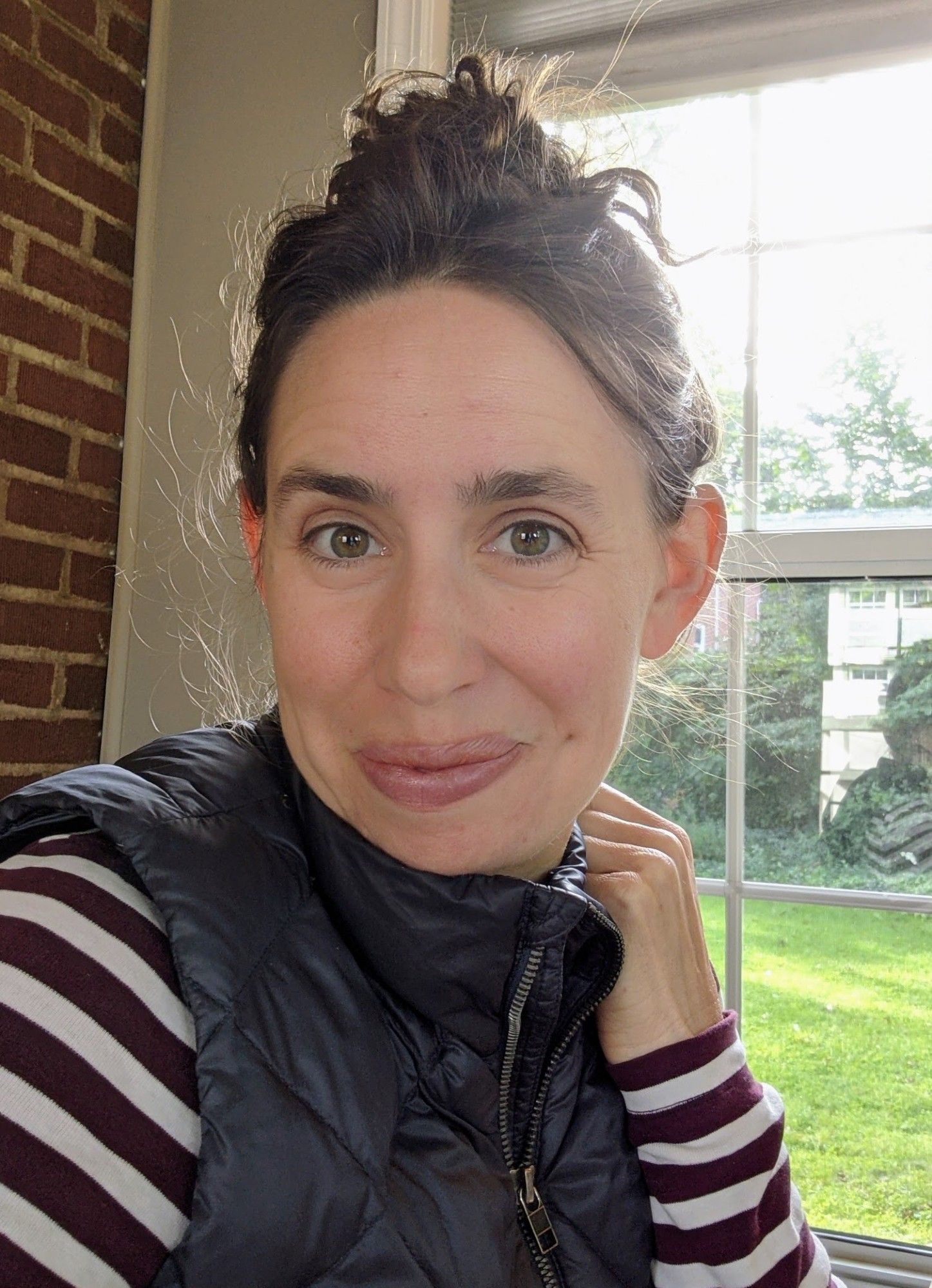 Hannah, a white woman with curly dark hair in a bun on top of her head, is giving a small, closed-mouth smile to the camera. Her left hand is near her chin. She's wearing a brown and white striped long-sleeved shirt with a blue puffer vest over it. There's a brick wall and a window looking out onto grass in the background.