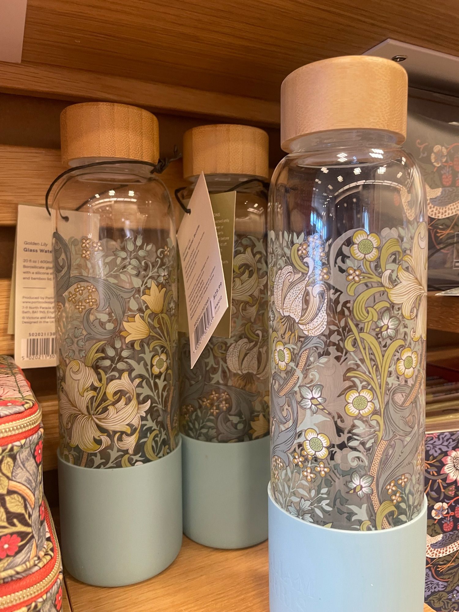 Three glass water bottles with scrolling green and blue botanical designs. The bottles have small light blue “sleeves” at the bottom and are sitting on a wooden shelf.