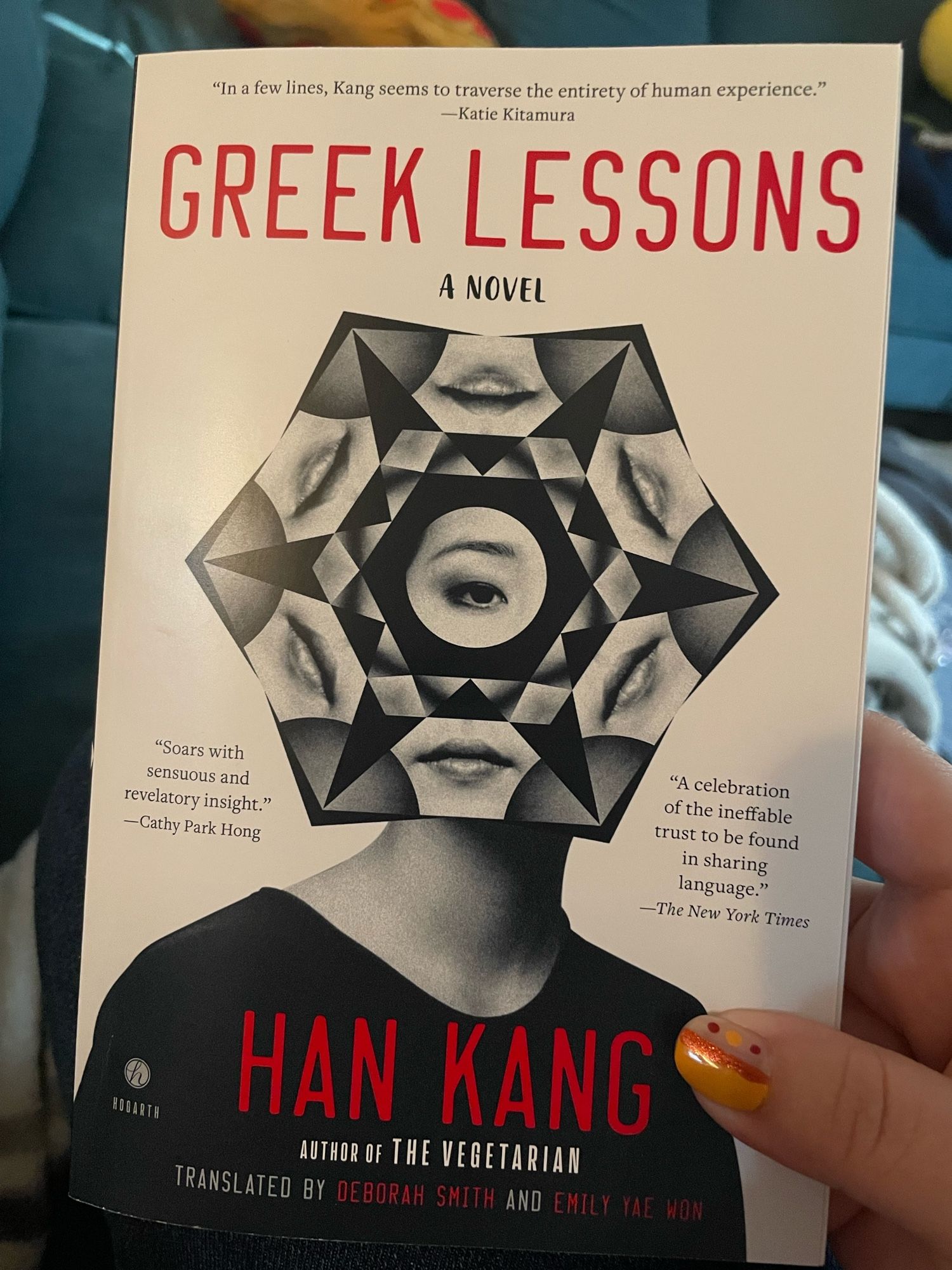 Cover of the paperback of Greek Lessons, by Han Kang. A figure in black and white has a kaleidoscopic hexagon over their face, with the shapes revealing many mouths and at the center, one eye. The text on the cover is black, white, and red, in sparse typefaces. 